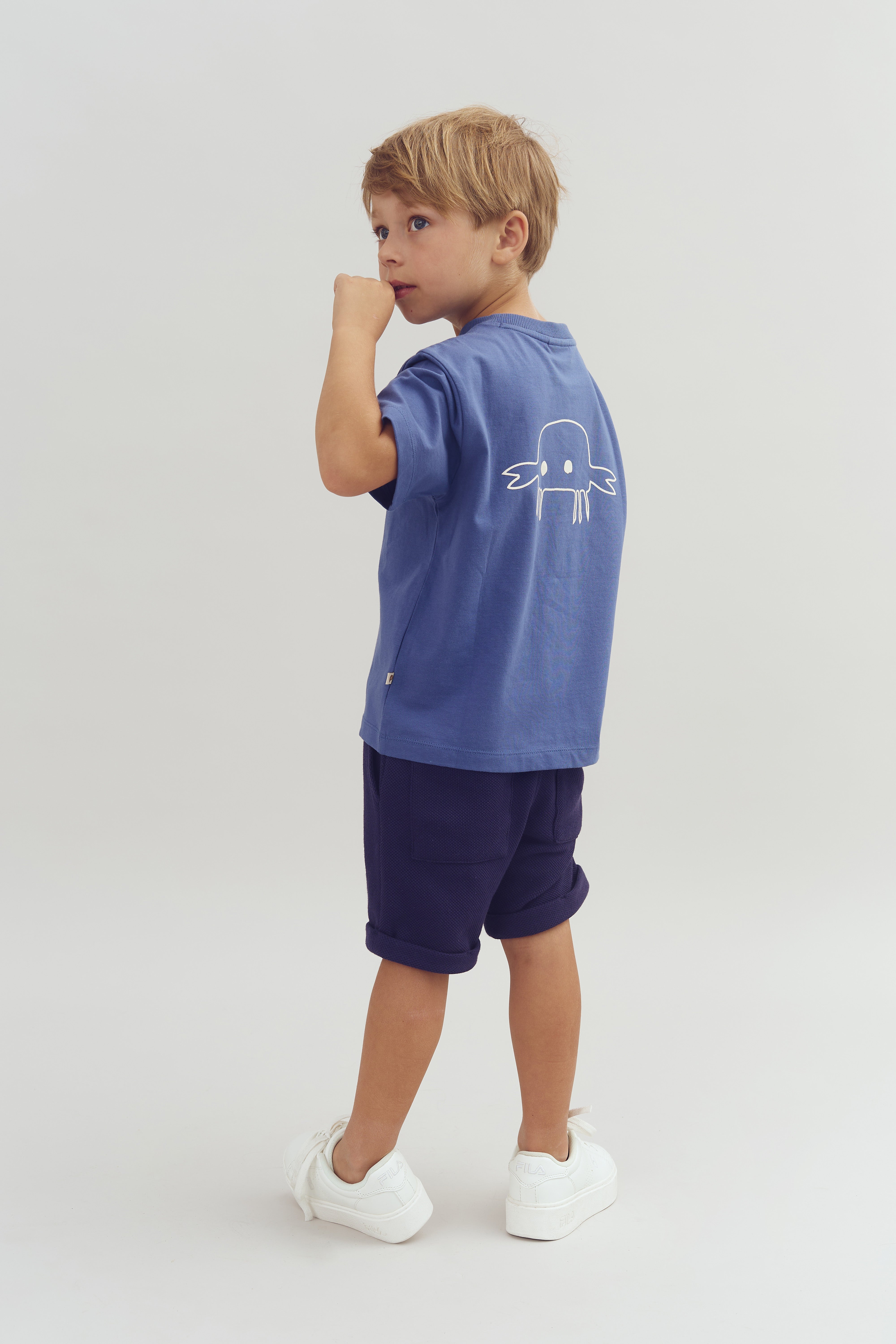 Jongen draagt blauw T-shirt met eenvoudige tekening op de rug, staat zijwaarts naar links kijkend en kauwt op zijn knokkel. Hij draagt ook donkerblauwe korte broek en witte sneakers.