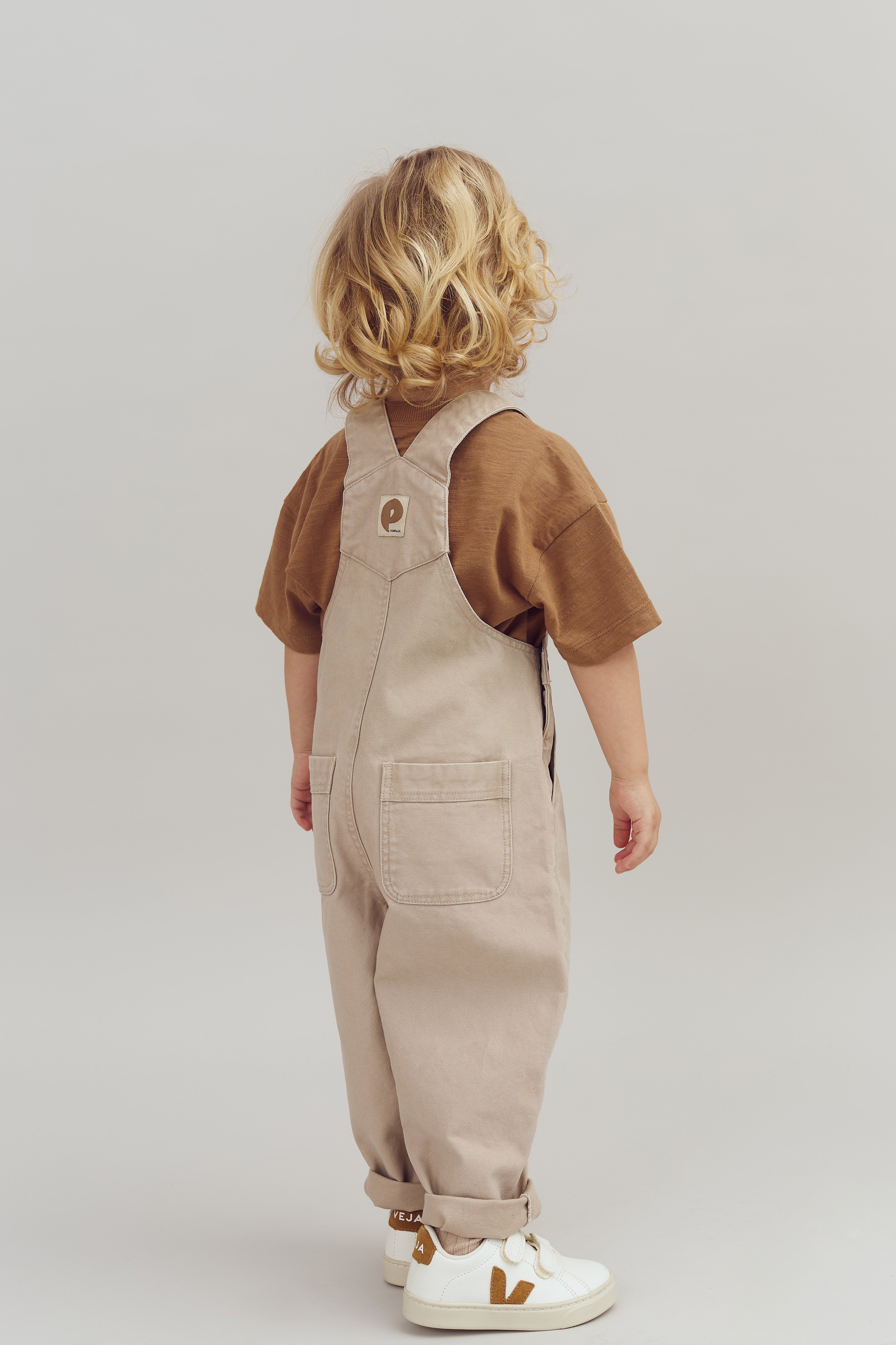 Een kind met golvend blond haar draagt een beige tuinbroek over een bruin shirt. Het staat op een vlakke, effen achtergrond. Op de schoenen staat "VEJA".