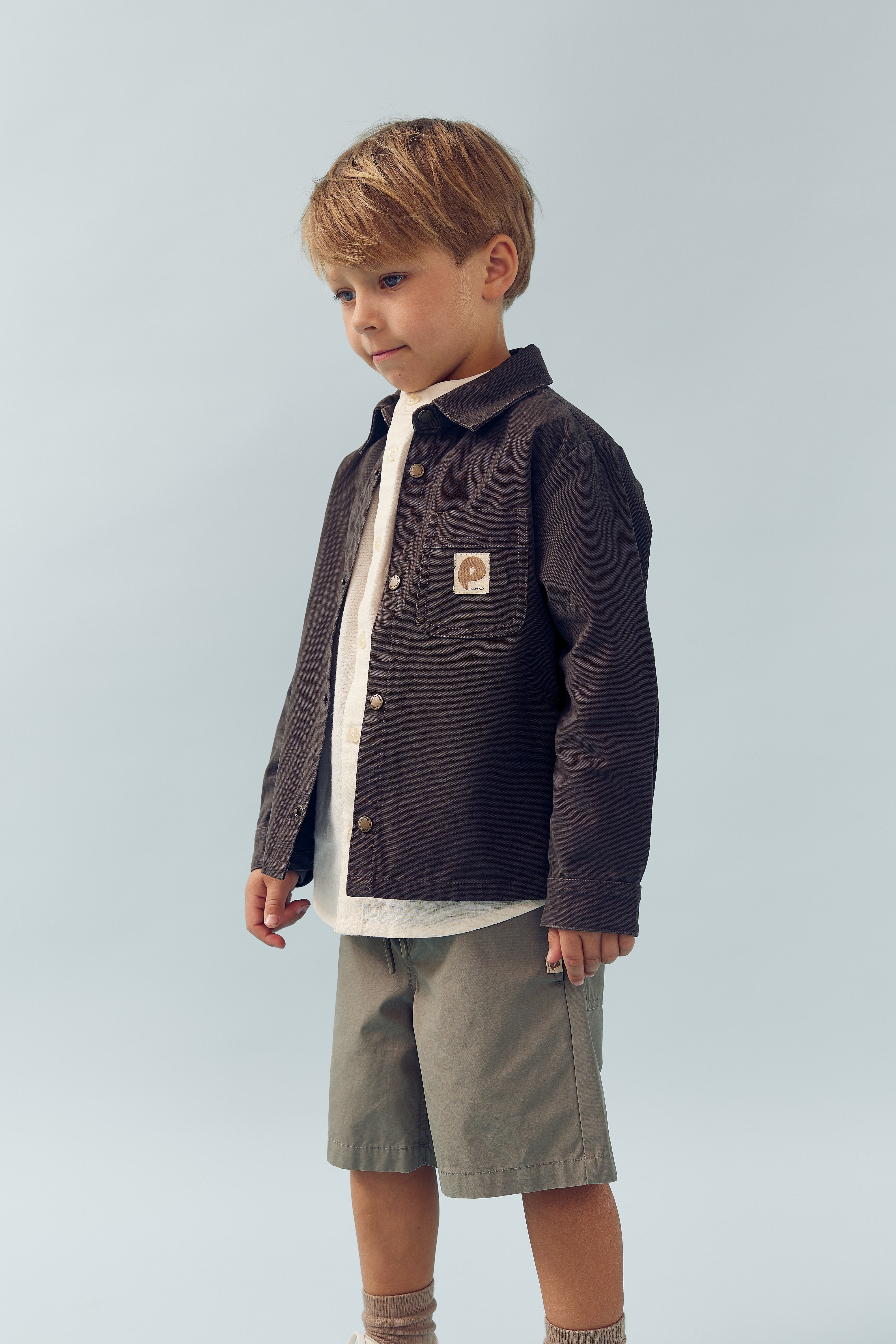 Een jongen draagt een donkerbruin jasje met een geborduurd label op de borst en groene shorts, staande voor een effen lichtblauwe achtergrond.