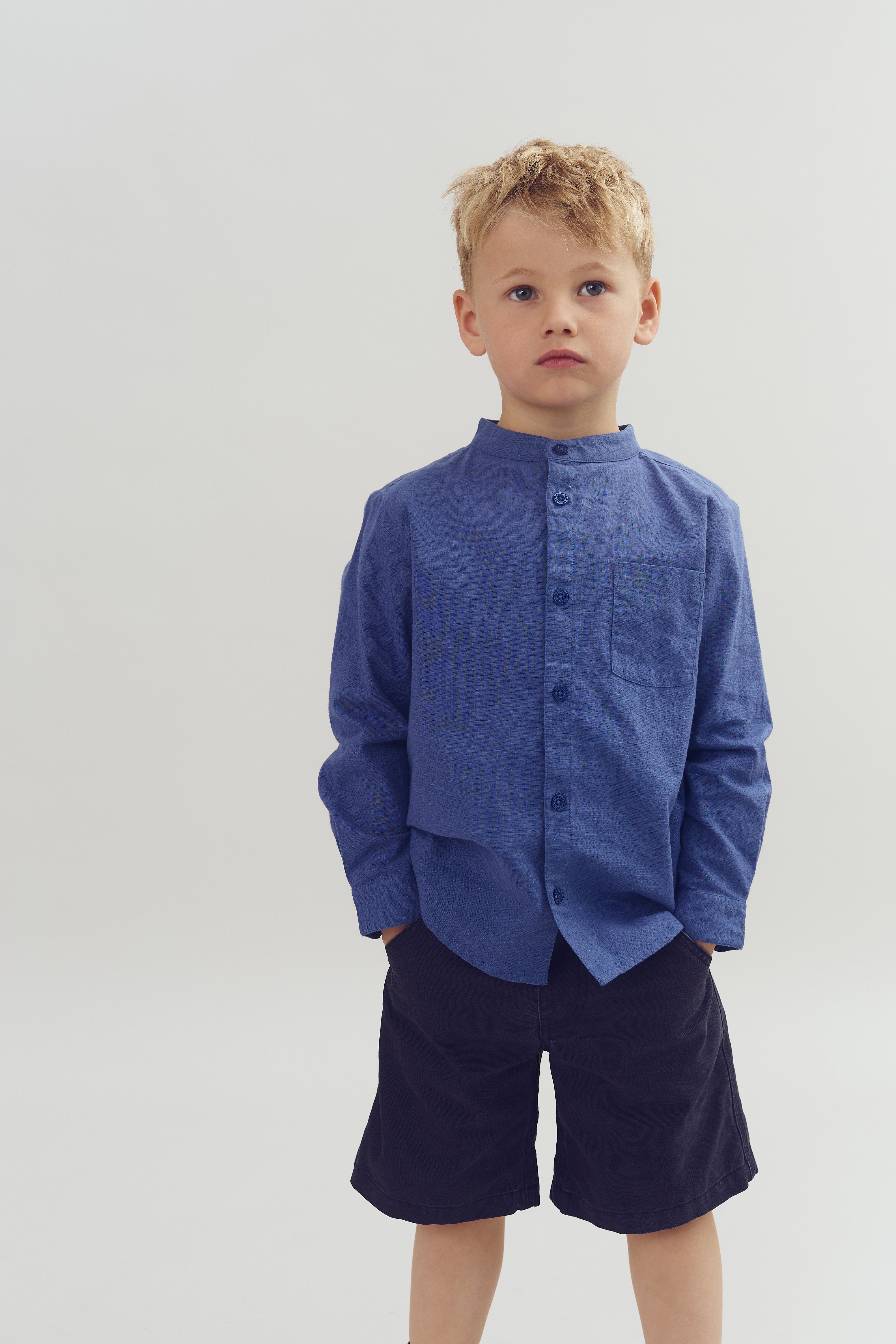 Jongen staat; draagt een blauw hemd met zwarte korte broek; handen in zakken; blanco achtergrond.