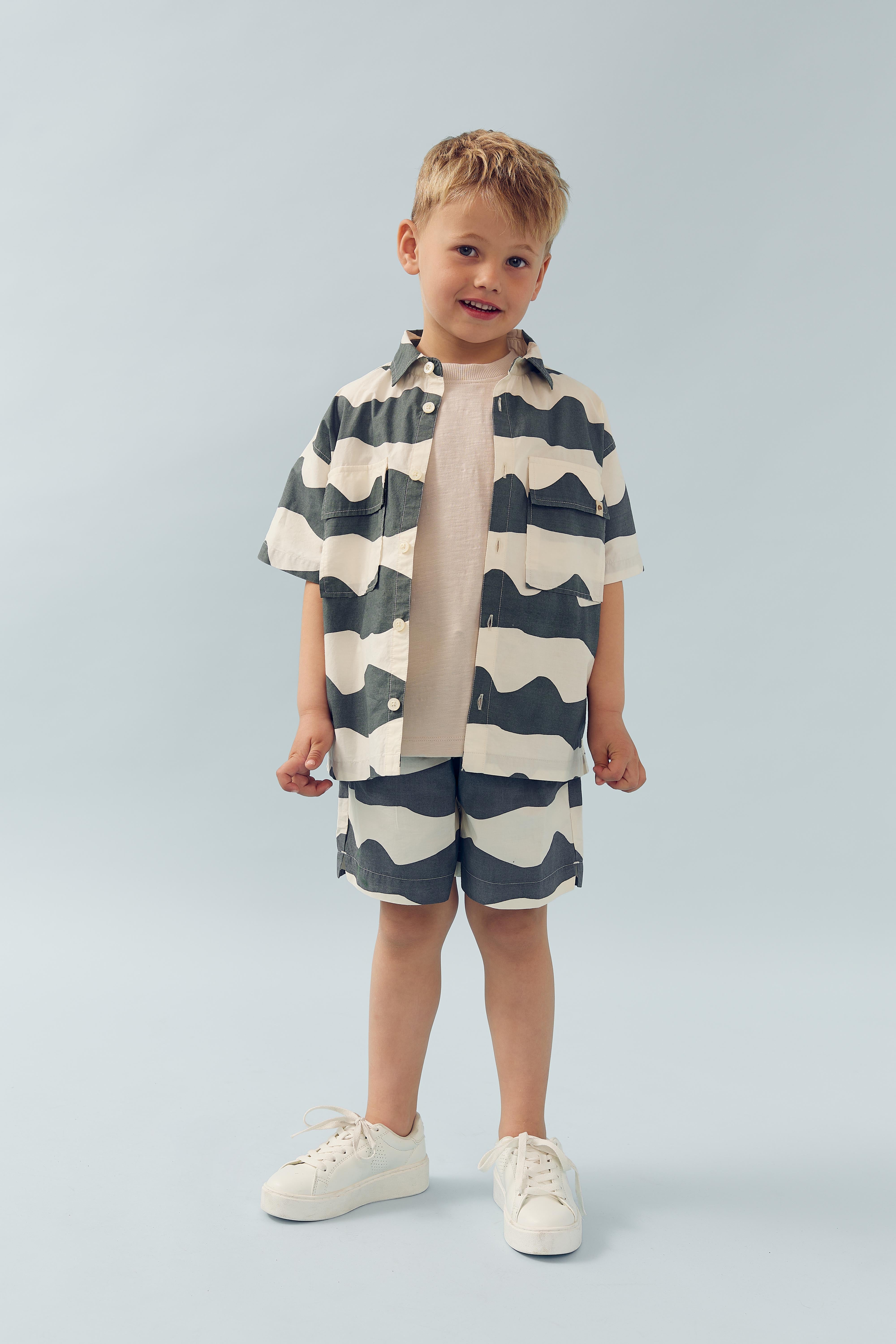 Een jongen staat en glimlacht, gekleed in een gestreepte korte broek en overhemd. Hij draagt witte schoenen. De achtergrond is effen lichtblauw.