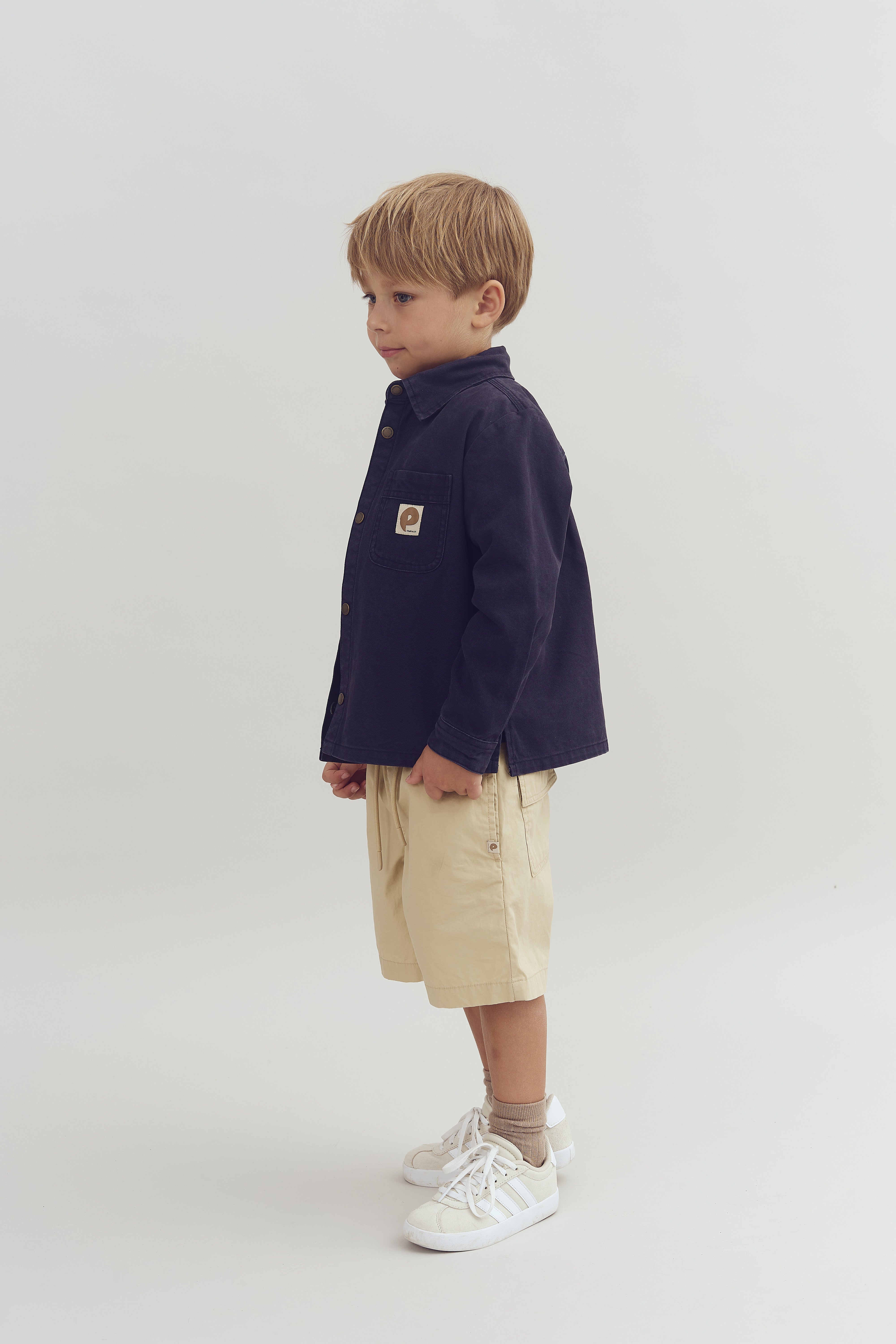 Kind loopt naar links, draagt een donkerblauw jasje met een embleem op de borstzak, beige shorts en witte sneakers. Neutrale, lichte achtergrond.