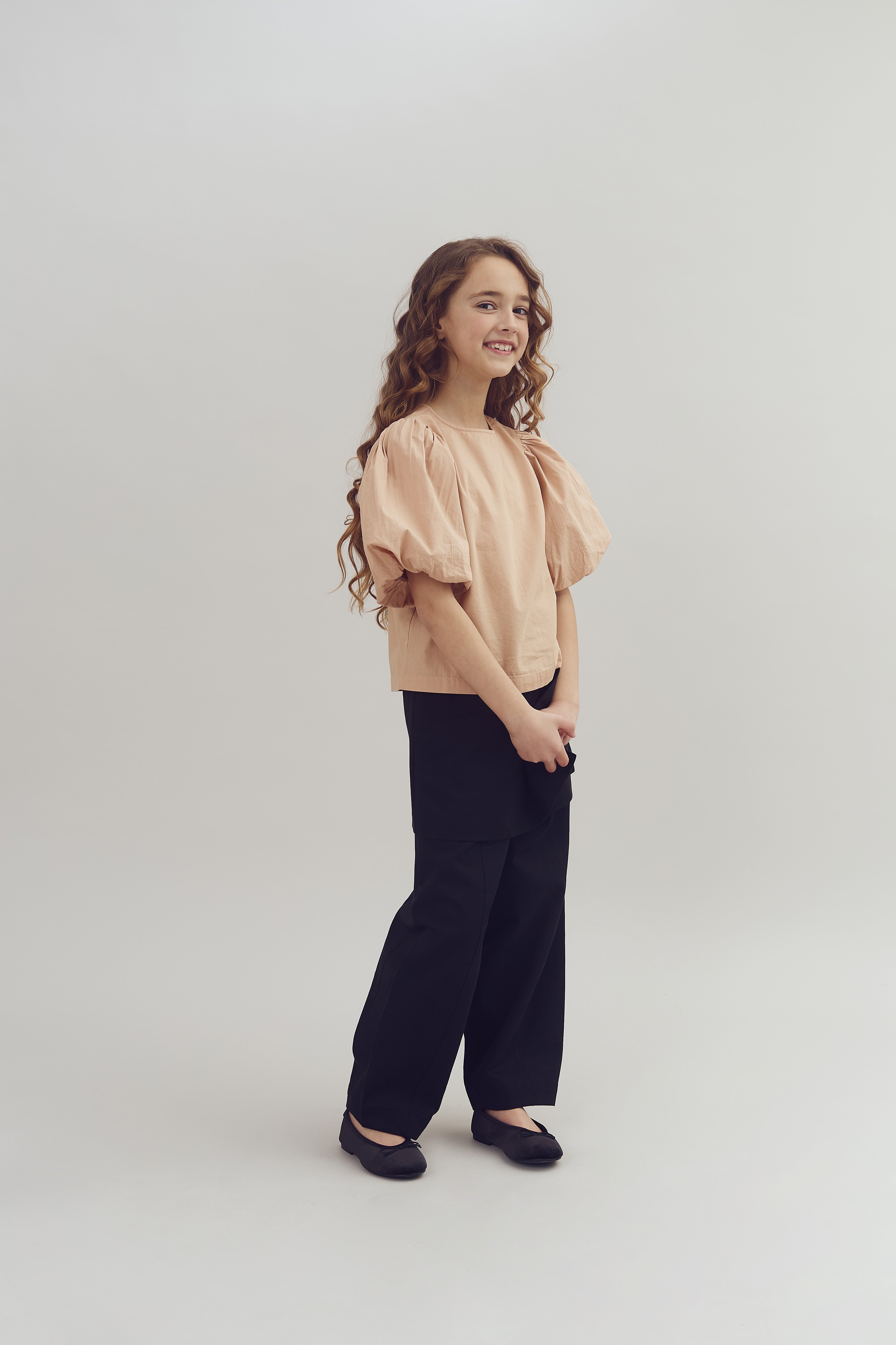 Meisje staat rechtop, draagt een beige blouse met pofmouwen en zwarte broek. Ze glimlacht en bevindt zich in een neutrale, lichte ruimte zonder tekst of opvallende elementen.