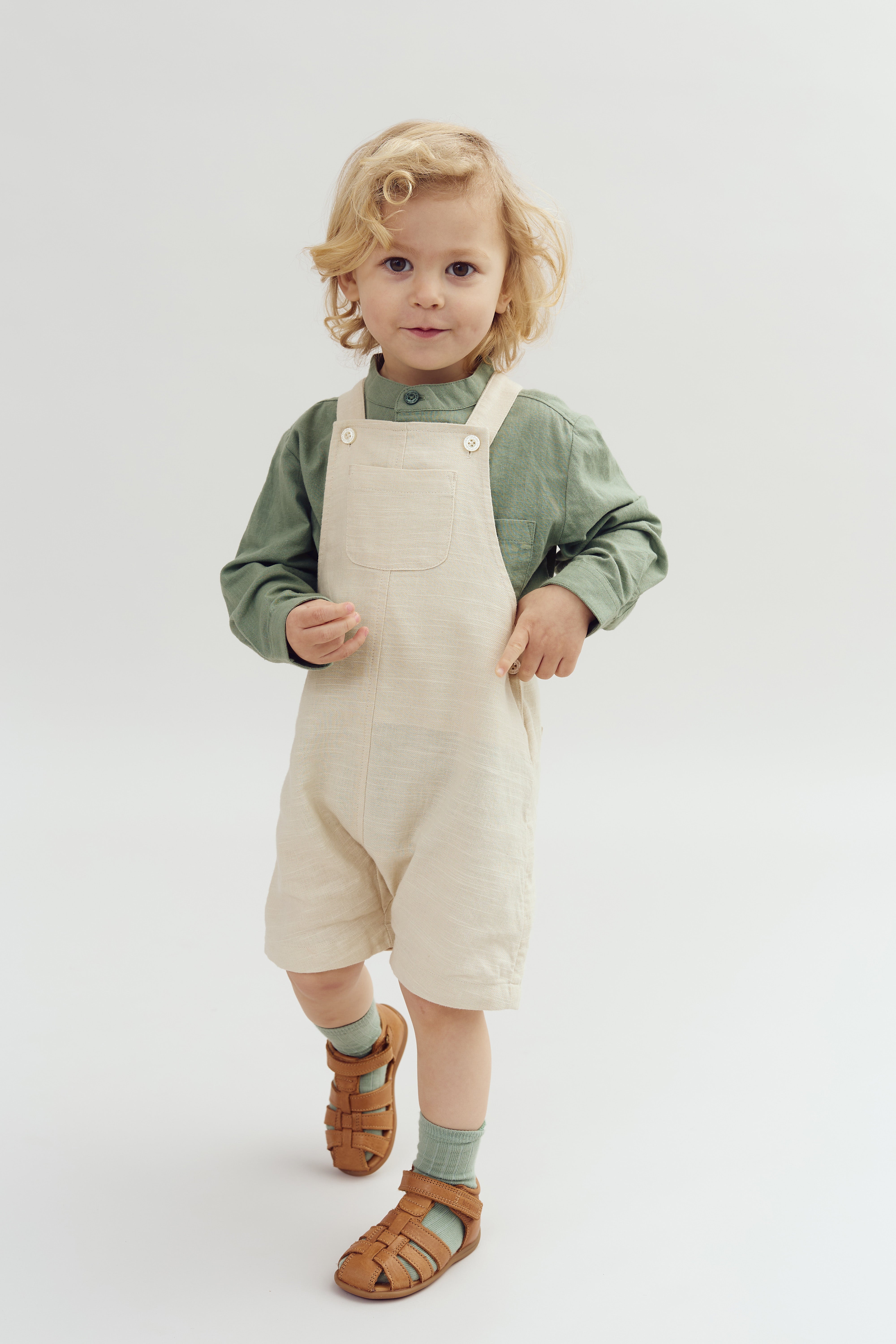 Jong kind met blond haar draagt groen shirt en beige tuinbroek, loopt op lichtgrijze achtergrond met bruine sandalen en groene sokken.
