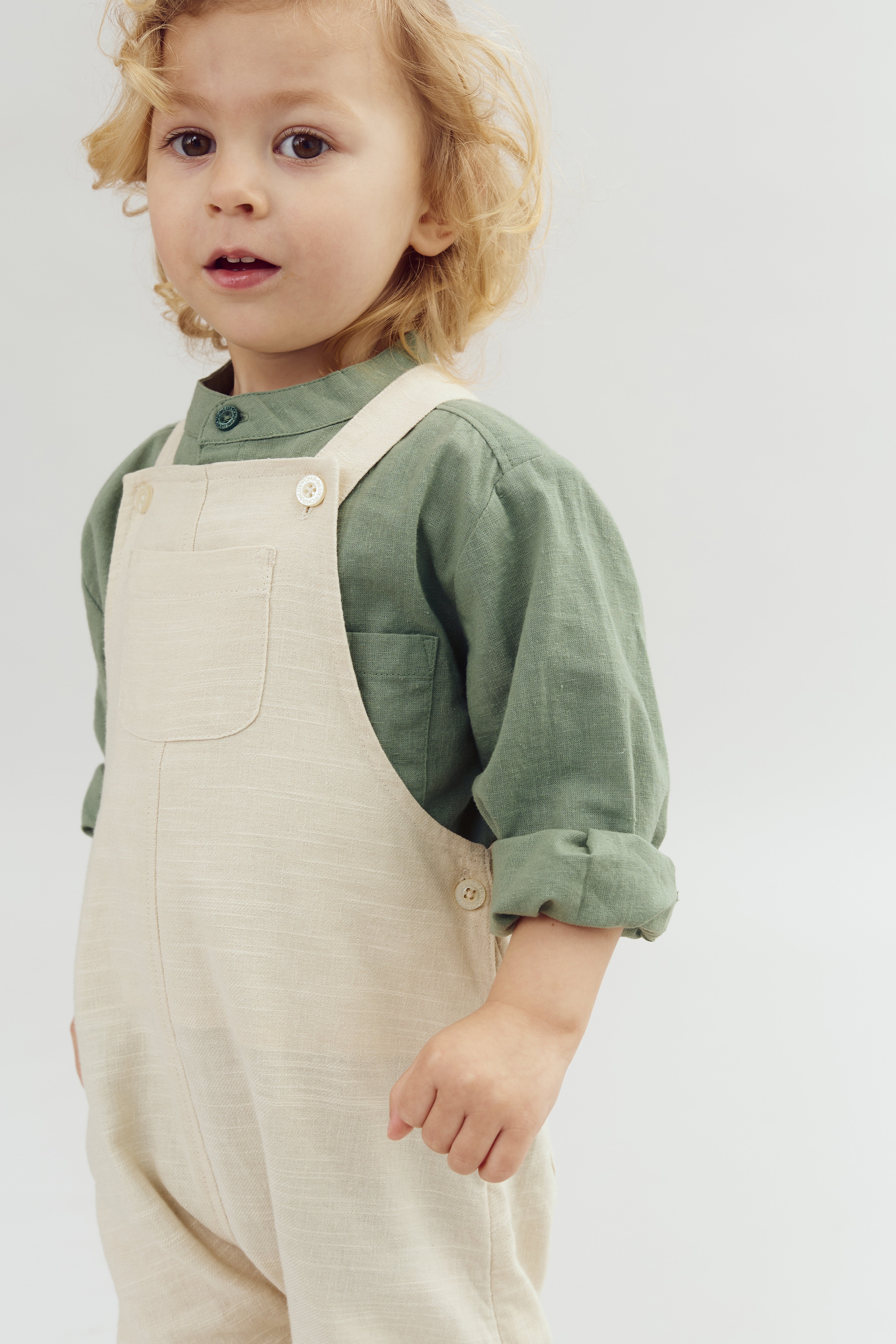 Een kind draagt een groen shirt en beige overall, staand in een neutrale, lichte omgeving, met licht krullend blond haar en een nieuwsgierige blik.
