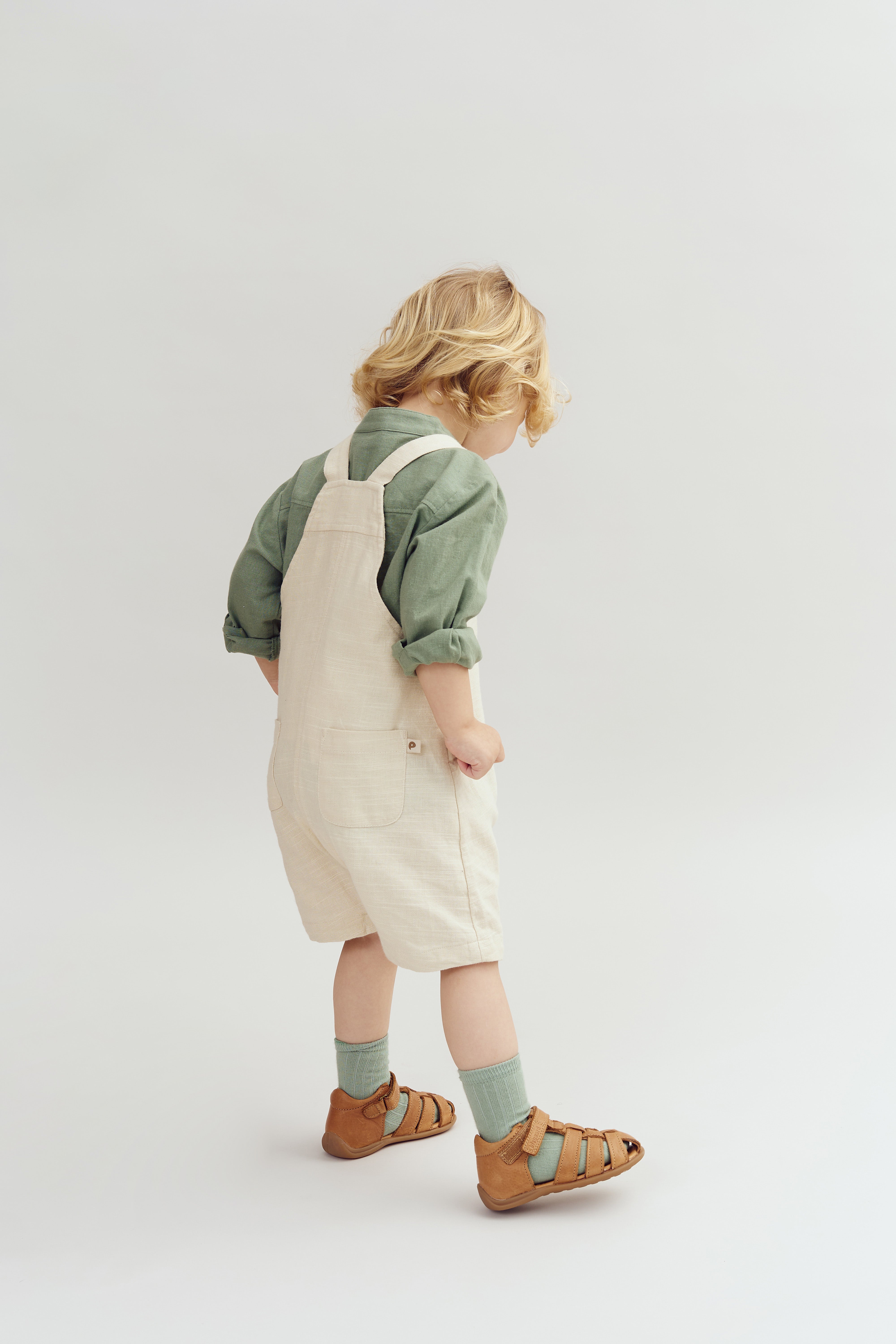 Een jong kind draagt een groene trui met een beige tuinbroek, wandelt in sandalen en sokken, tegen een neutrale, lichtgrijze achtergrond.