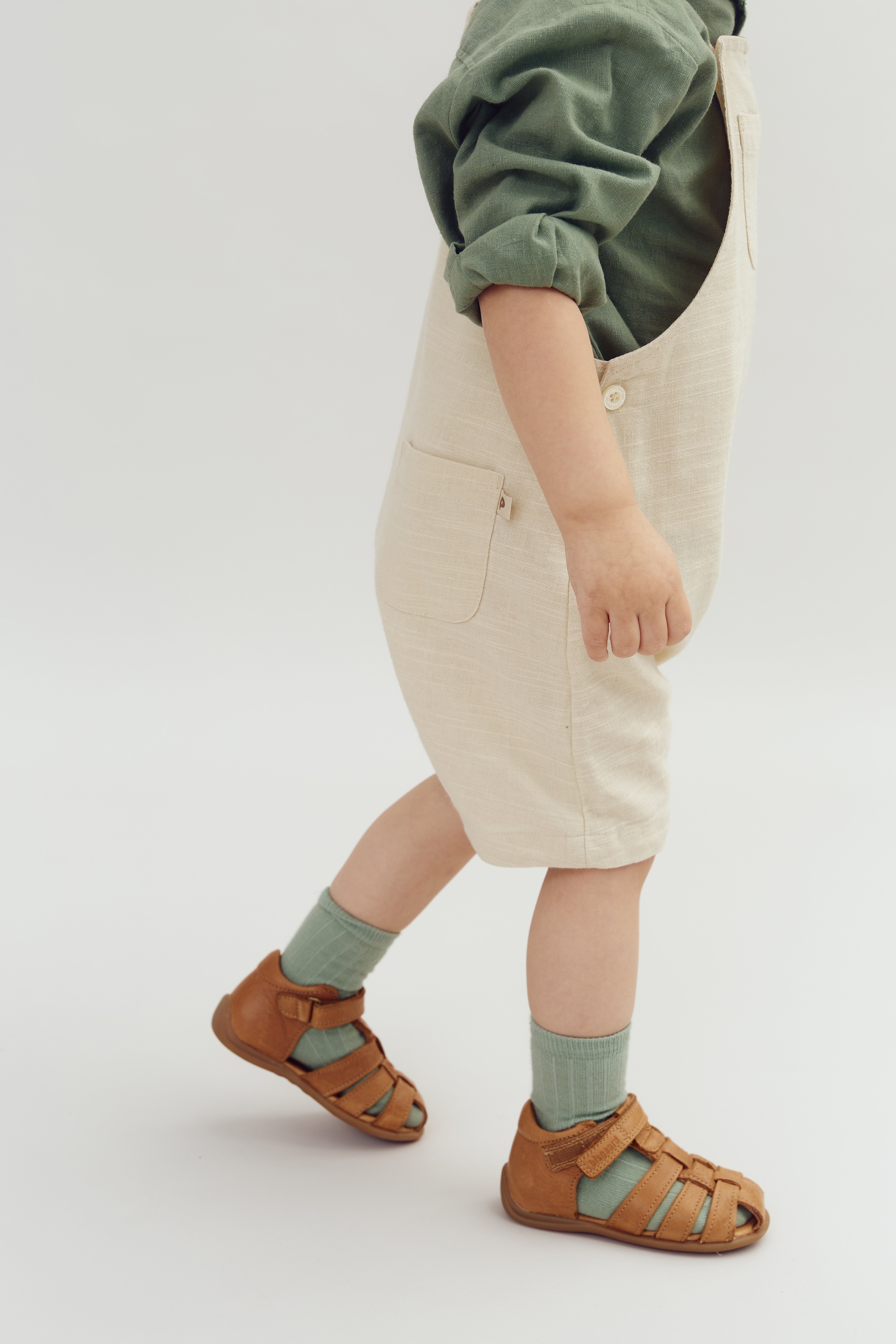 Kind draagt beige tuinbroek en groene trui, loopt rustig. Omgeving is een witte, lege achtergrond. Het kind draagt bruine sandalen met groene sokken.
