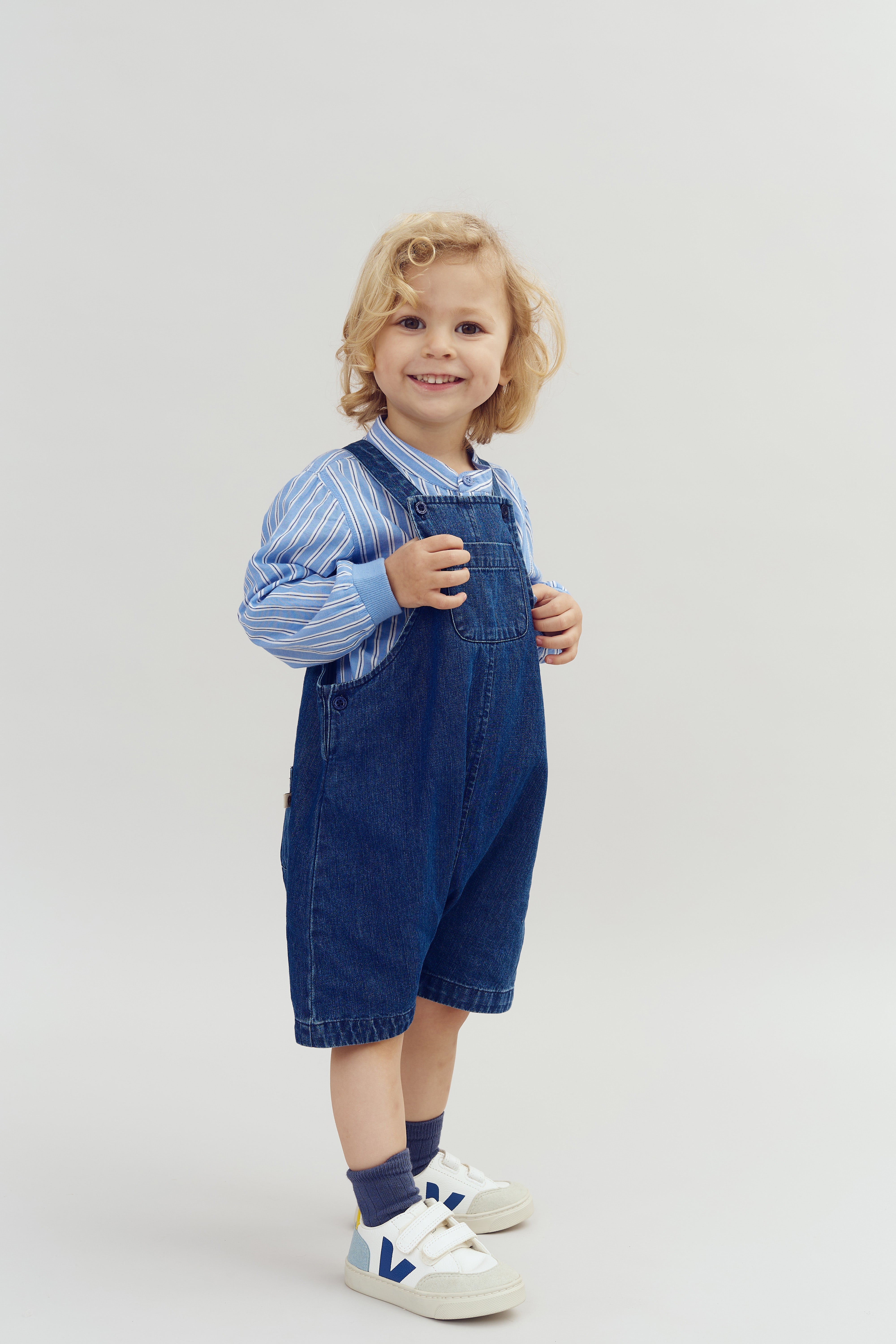 Een kind glimlacht, draagt een blauwe gestreepte blouse en een denim tuinbroek, in een lichte, neutrale ruimte, balancerend op witte sneakers met blauwe accenten.