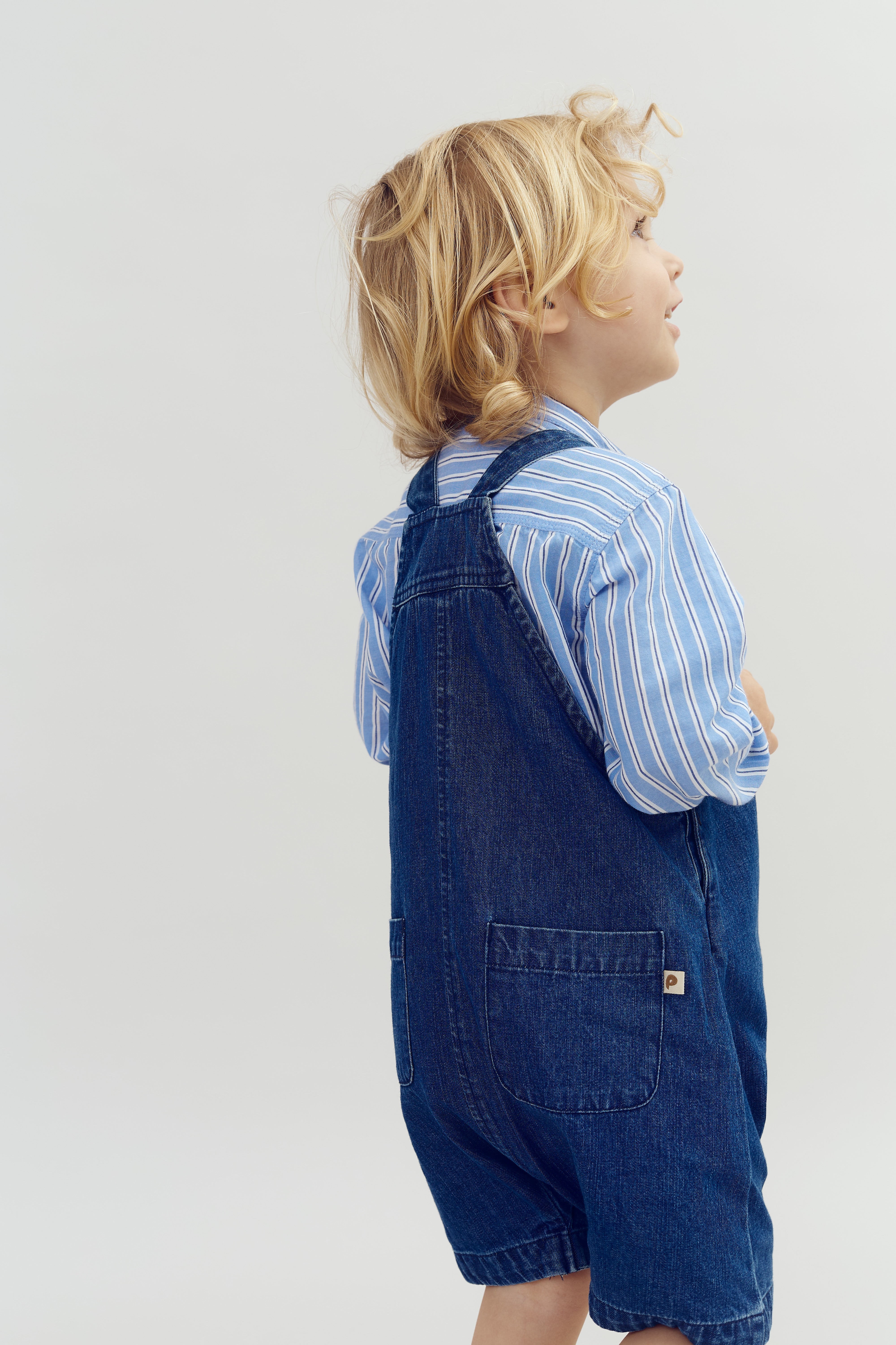 Een kind met blond haar draagt een blauw-wit gestreepte blouse en een jeans tuinbroek, staande zijwaarts tegen een neutrale, lichte achtergrond.