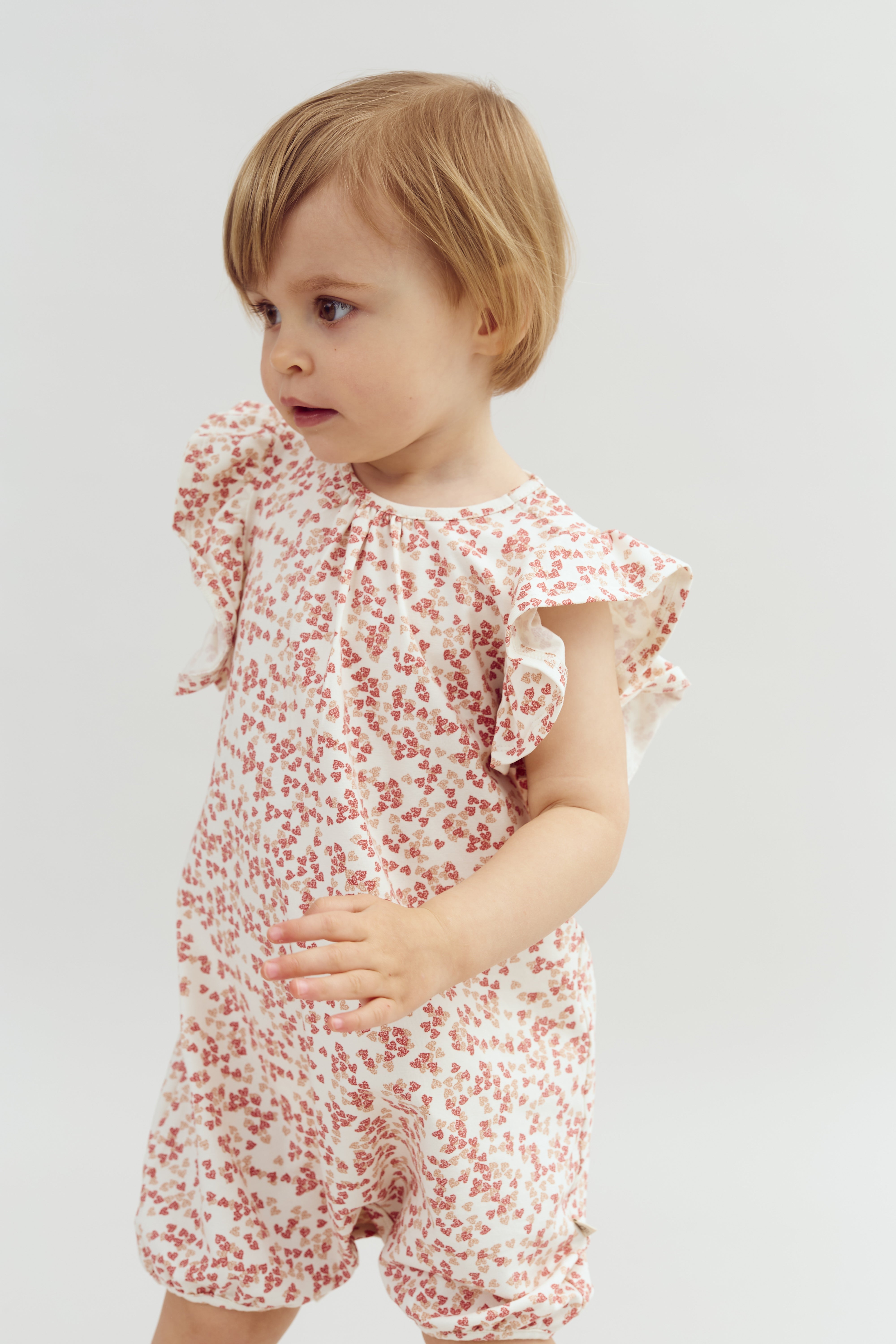 Peuter draagt een witte romper met rode bloemen. Het kind kijkt naar links, met haar los blond haar. De achtergrond is een effen, lichtgekleurde omgeving.