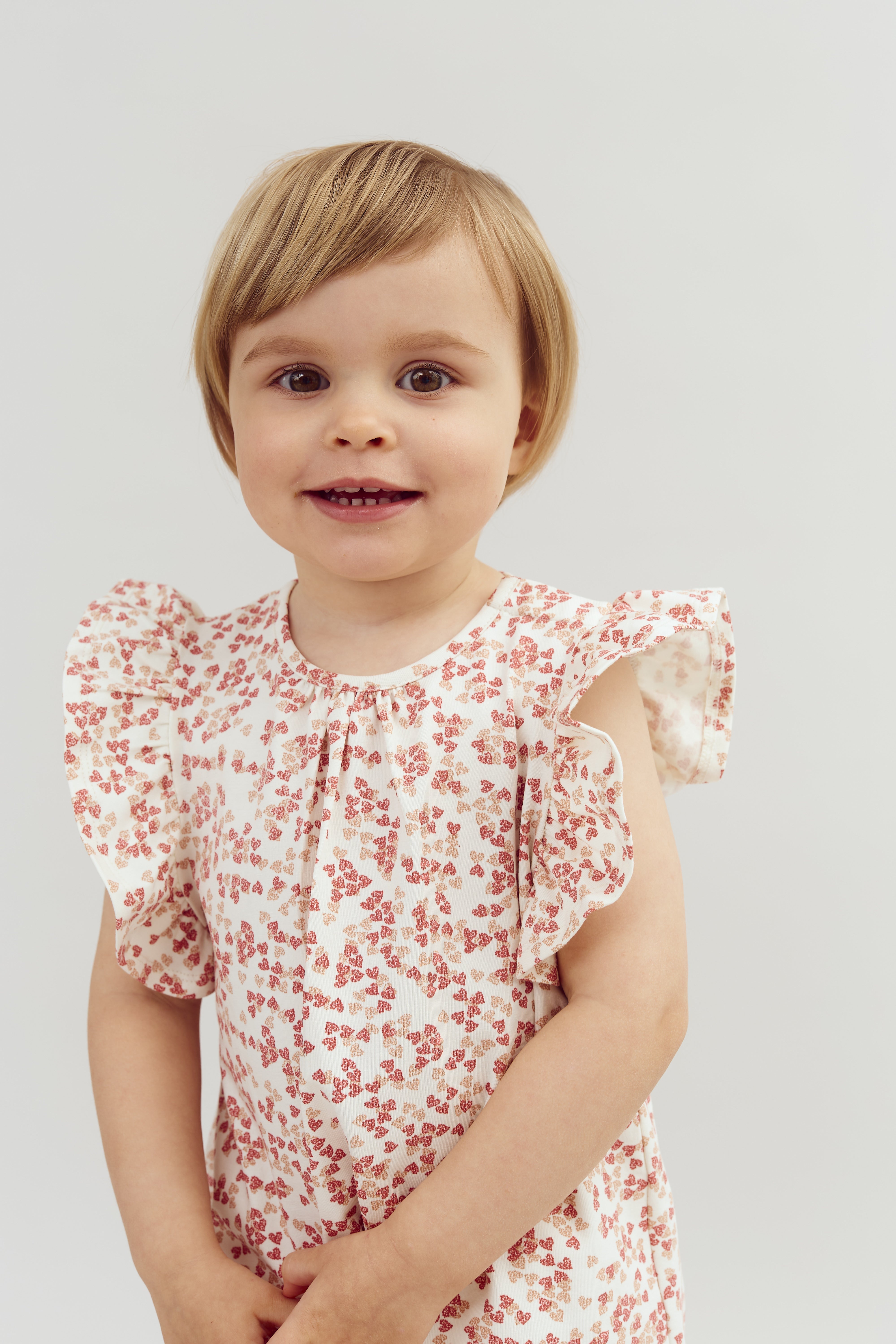 Een klein kind glimlacht, draagt een wit jurkje met roze bloemen en staat in een eenvoudige, lichte achtergrond.