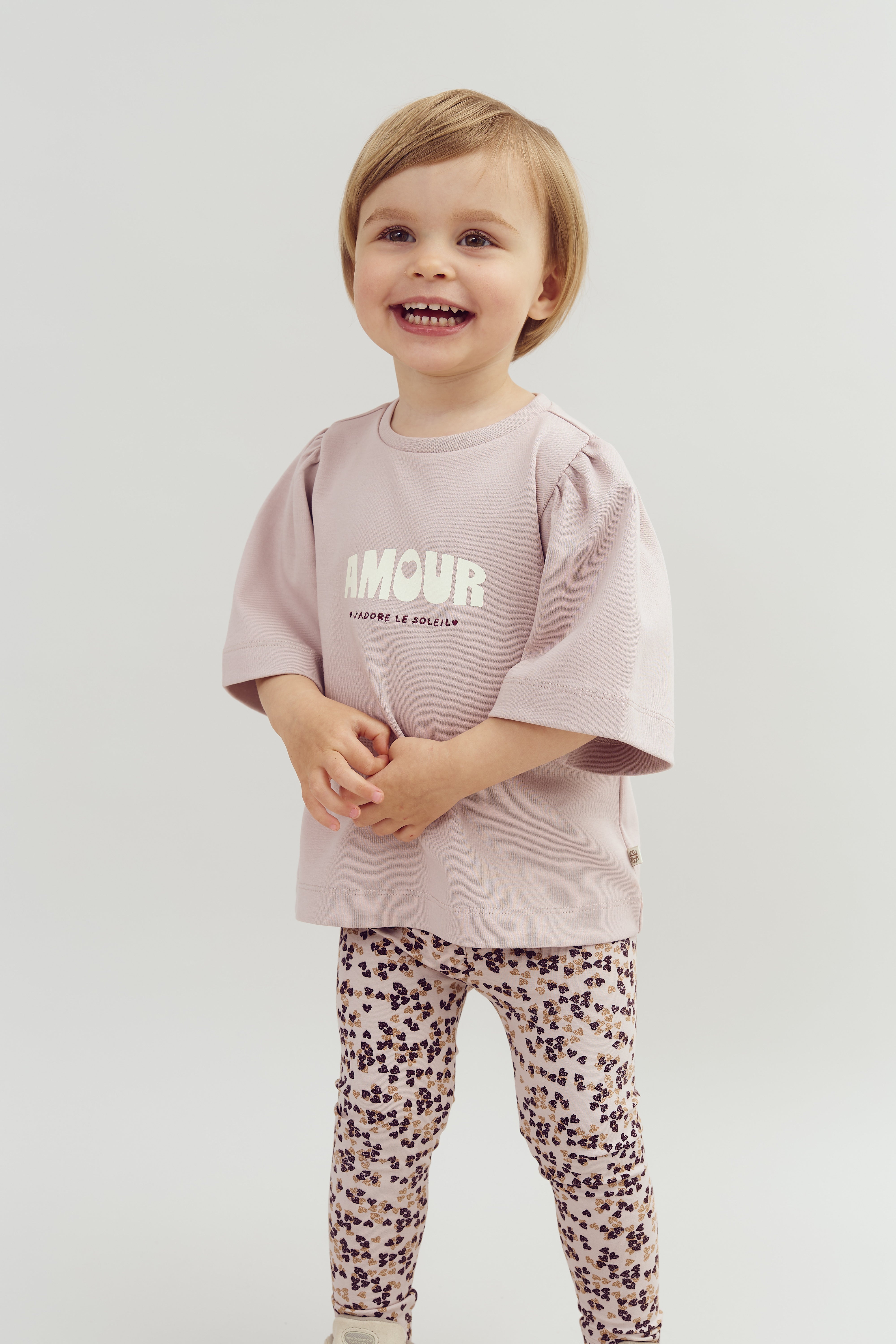 Een jong kind lacht, gekleed in een lichtroze shirt met de tekst "AMOUR J'ADORE LE SOLEIL" en een legging met bloemenpatroon, tegen een neutrale achtergrond.