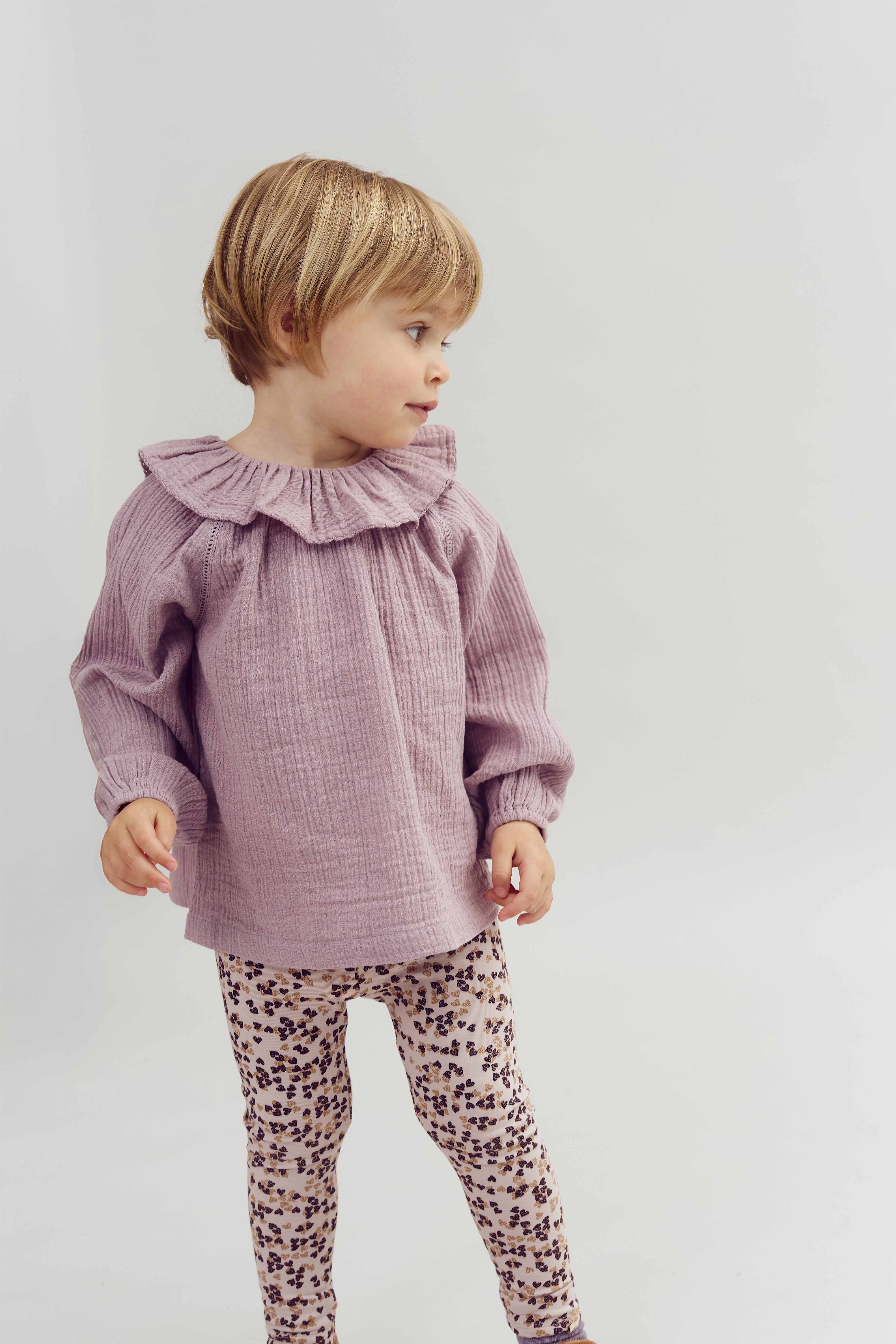 Een kind draagt een lila blouse met een rucheskraag en bedrukte legging. Het kijkt opzij tegen een neutrale, lichte achtergrond.