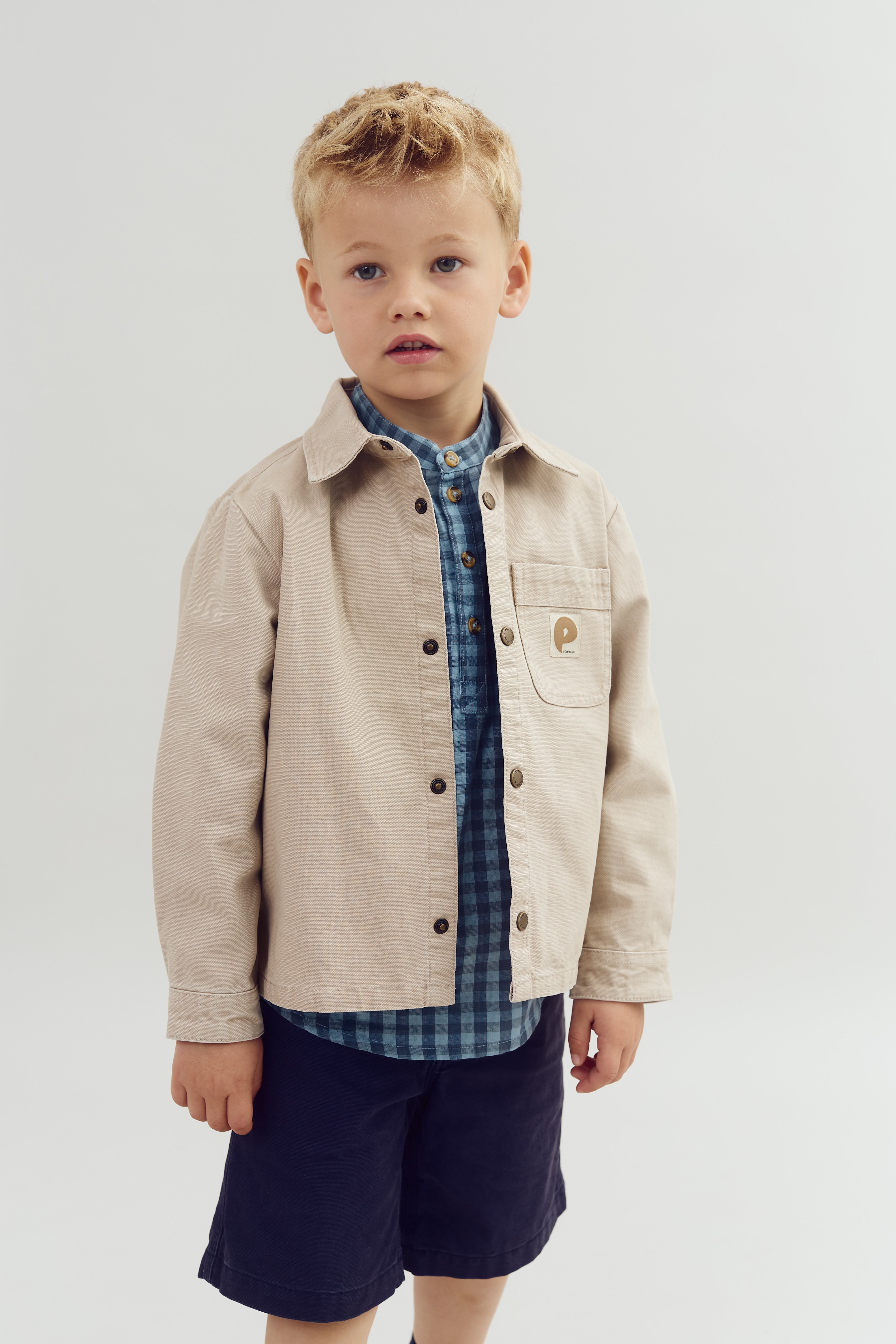Jongen draagt een beige jas met een P-logo op de borstzak en een blauw geruite blouse. Hij staat in een neutrale, lichte omgeving.