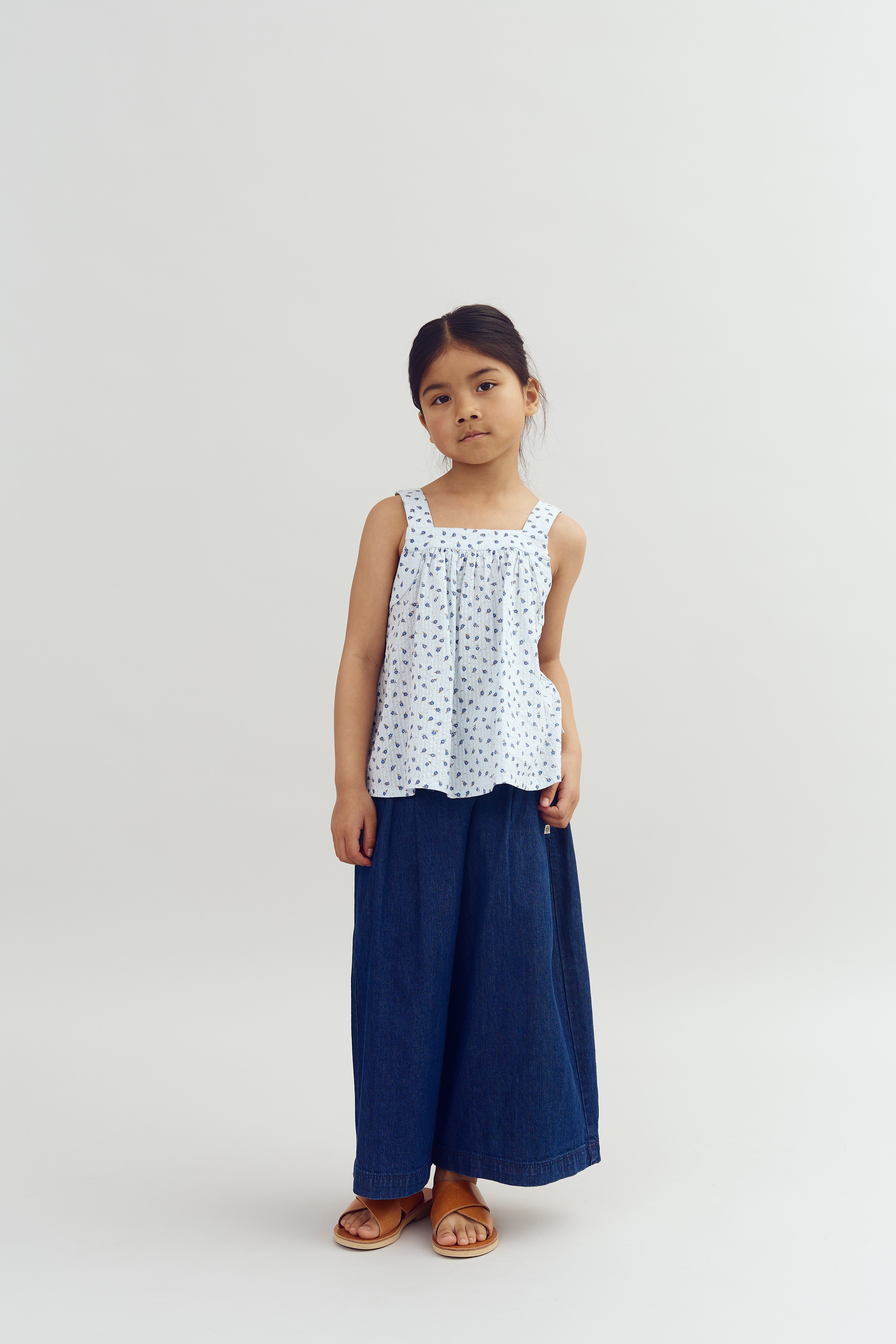 Een jong meisje staat rechtop, draagt een witte top met patroon en een wijde blauwe broek. Ze bevindt zich in een neutrale, lichte ruimte.