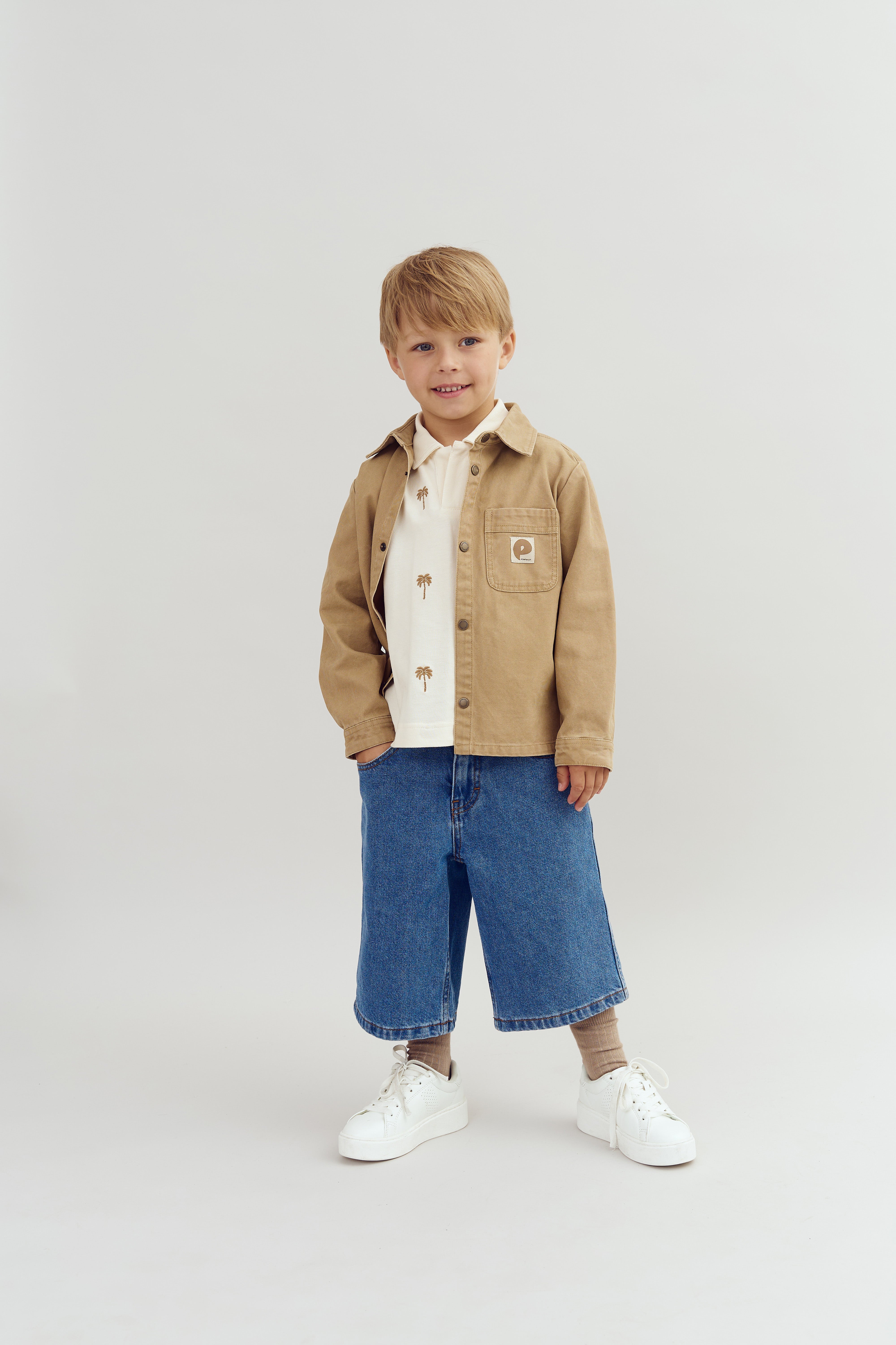 Kind staat lachend, draagt een beige jas en een wit shirt met palmbomen, blauwe spijkerbroek en witte schoenen, tegen een eenvoudige, lichte achtergrond.
