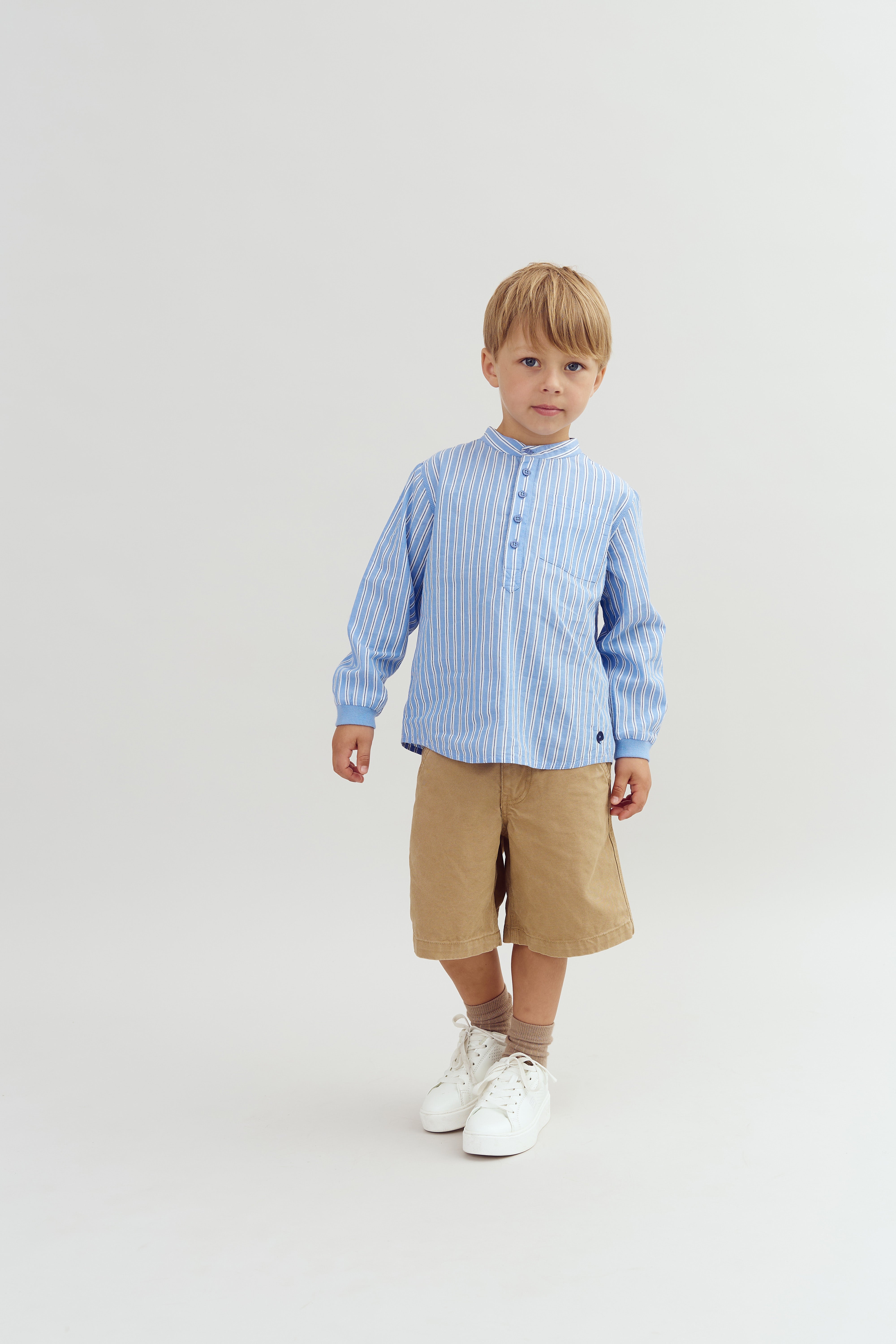 Een jong kind staat in een studio. Hij draagt een lichtblauw gestreept overhemd, beige shorts en witte schoenen. De achtergrond is effen en helder verlicht.