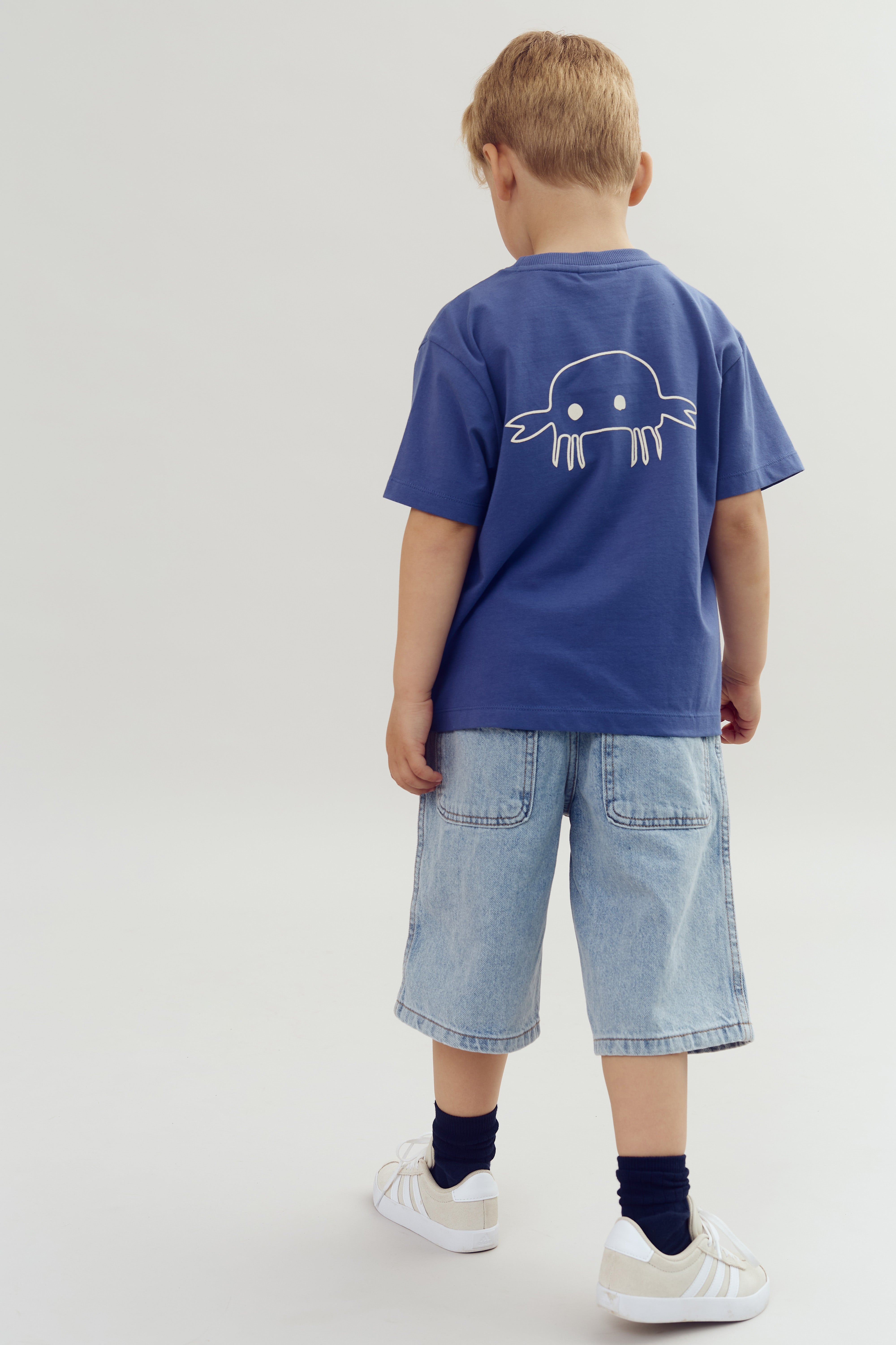 Een kind draagt een blauw T-shirt met een witte tekening van een krab, gecombineerd met een lichte spijkerbroek en witte sneakers, staand tegen een neutrale achtergrond.