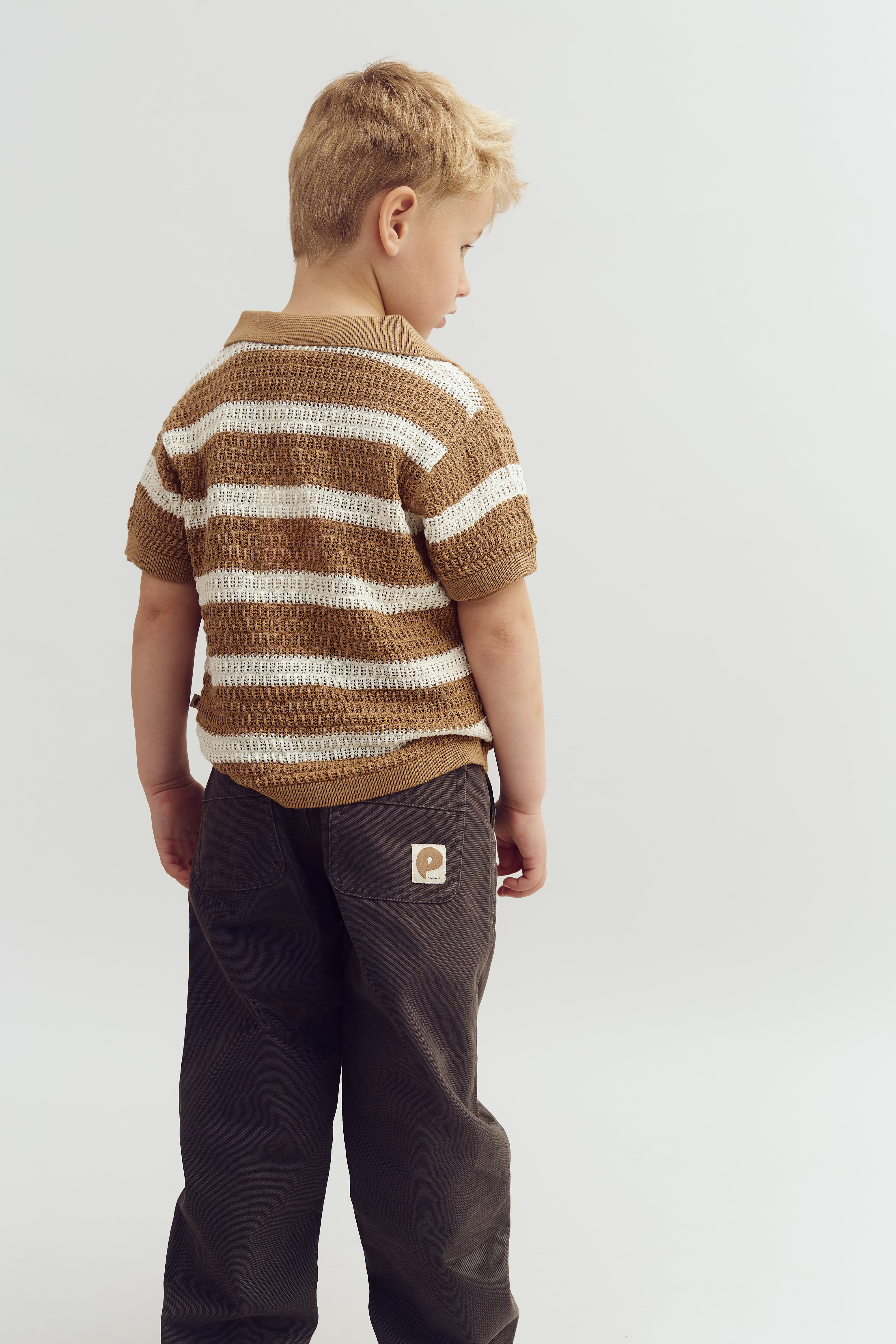Een jongetje staat met zijn rug naar de camera, gekleed in een bruin-wit gestreept shirt en donkere broek, binnen een minimalistische, lichte studio.