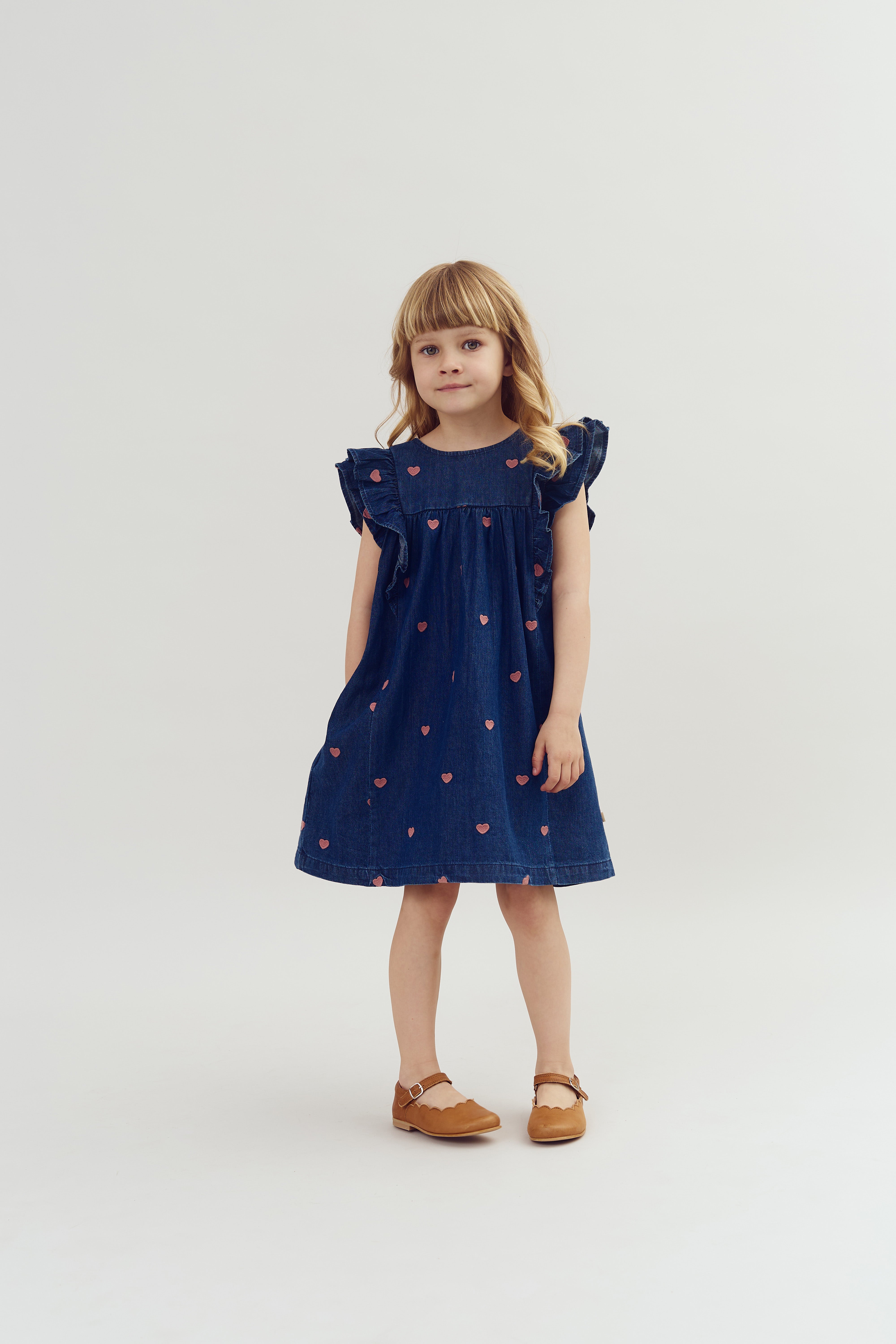 Een jong meisje draagt een blauwe jurk met roze hartjes en bruine schoenen. Ze staat rechtop in een eenvoudige, witte studio-omgeving.