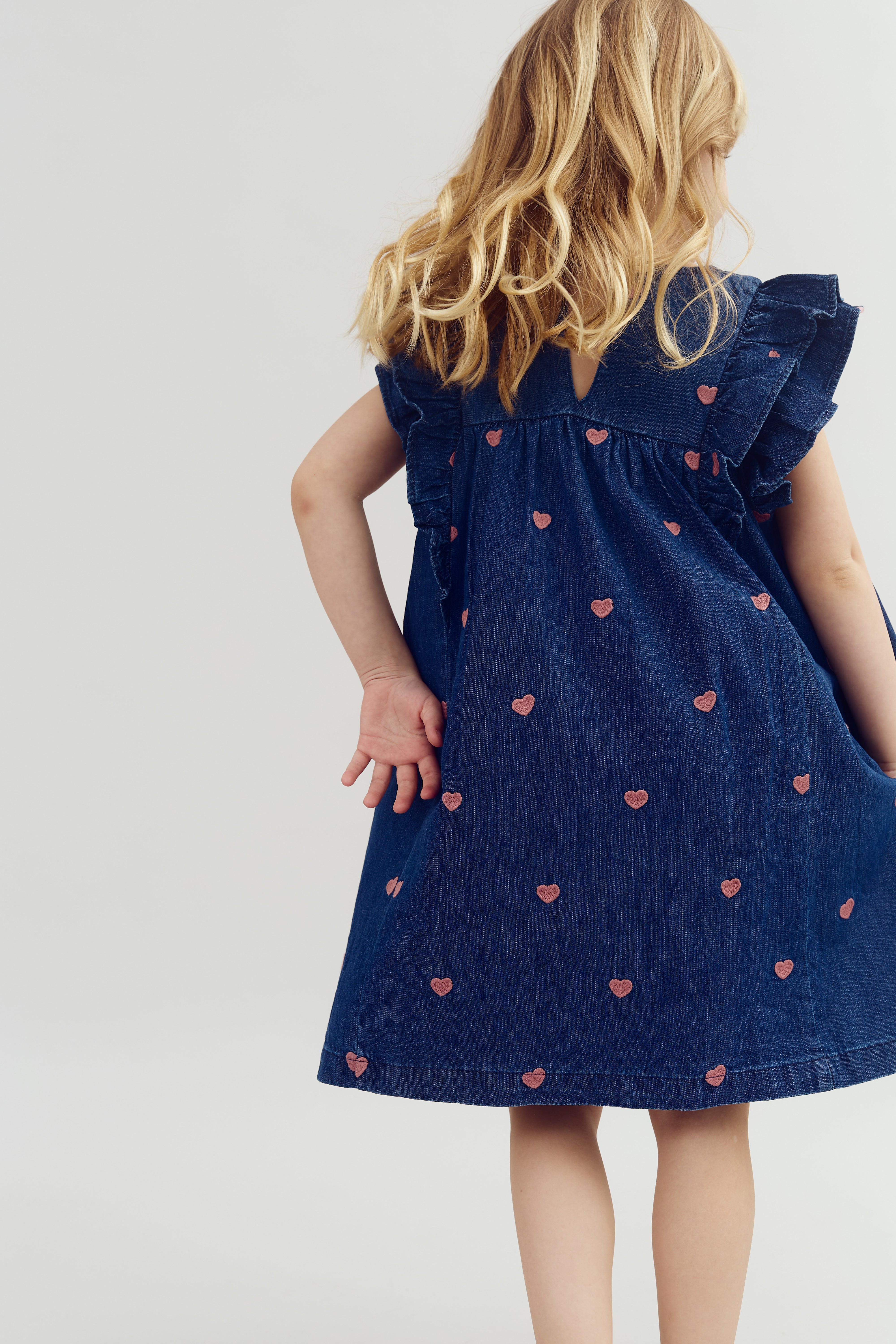 Een kind draagt een donkerblauwe jurk met roze hartjesmotief en staat met haar rug naar de camera. Ze is omringd door een neutrale, witte achtergrond.