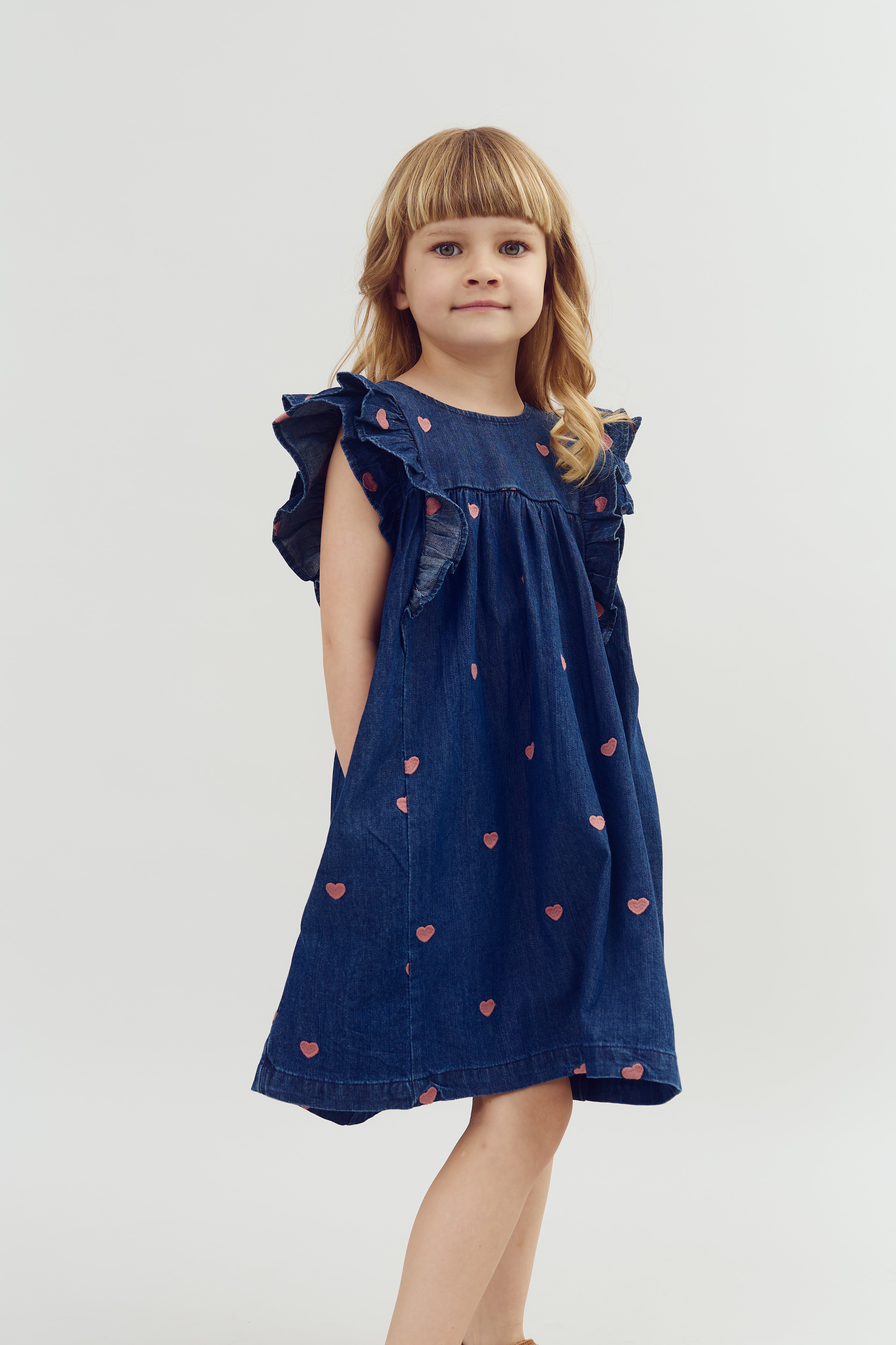 Een kind draagt een donkerblauwe jurk met roze hartjes en rouches. Het staat tegen een witte achtergrond.