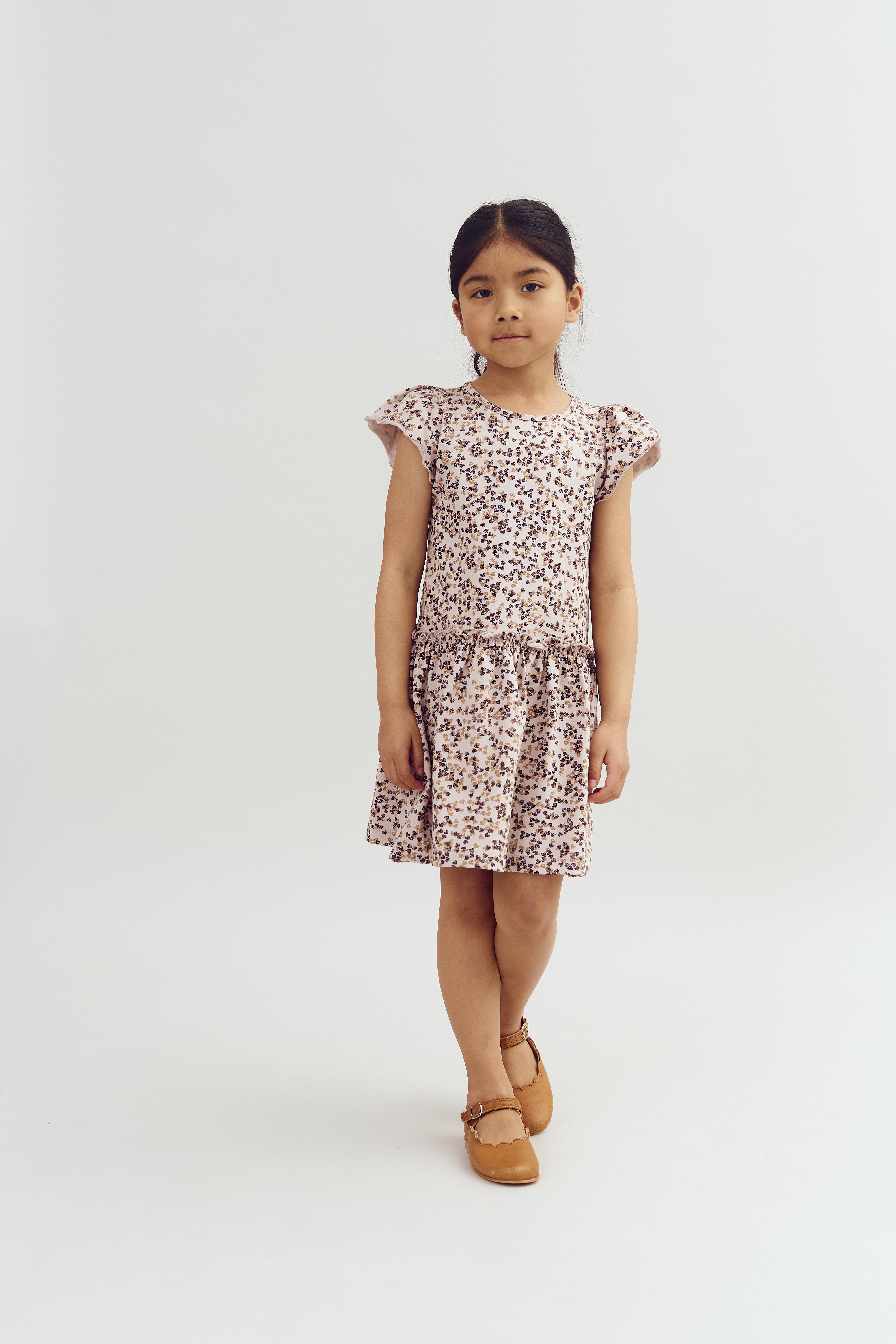 A young girl stands confidently, wearing a floral dress and brown shoes, against a plain, white background.