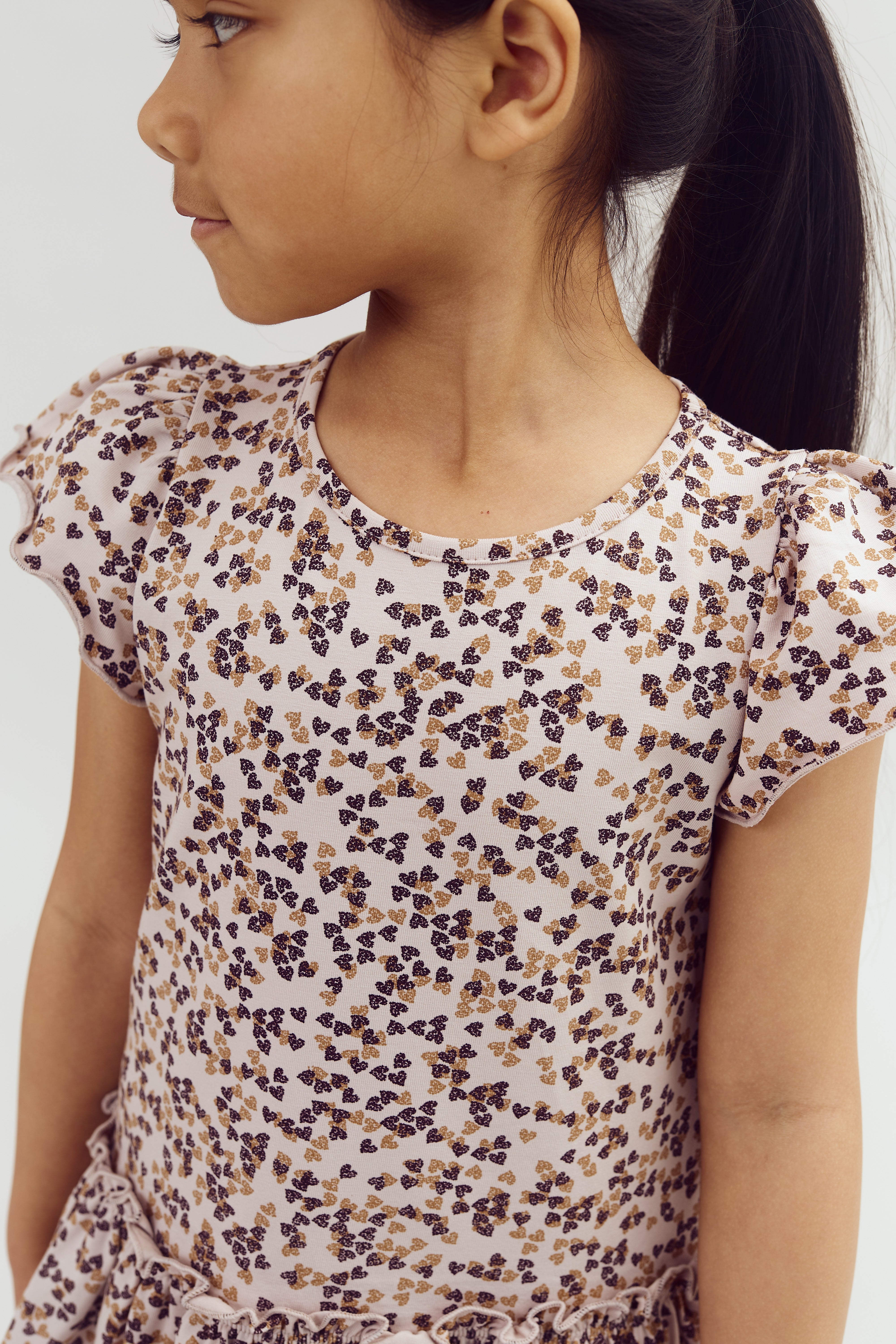 A young girl looks to the side, wearing a patterned dress with brown and black floral designs against a light background. Her dark hair is tied back, set against a plain white backdrop.