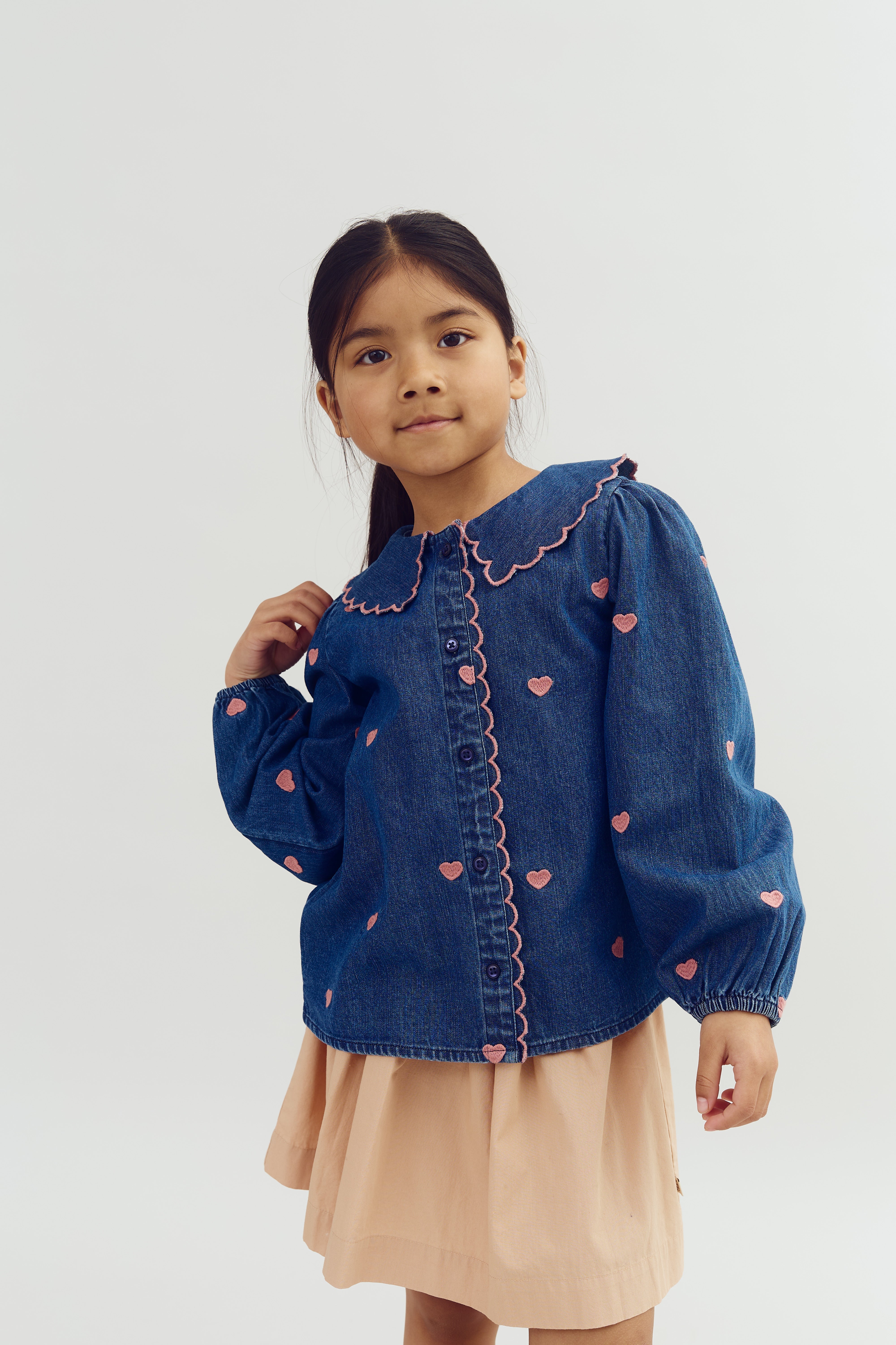 Meisje, gekleed in een blauwe blouse met roze hartjes en een beige rok, poseert terwijl zij haar haar aanraakt tegen een neutrale, lichte achtergrond.