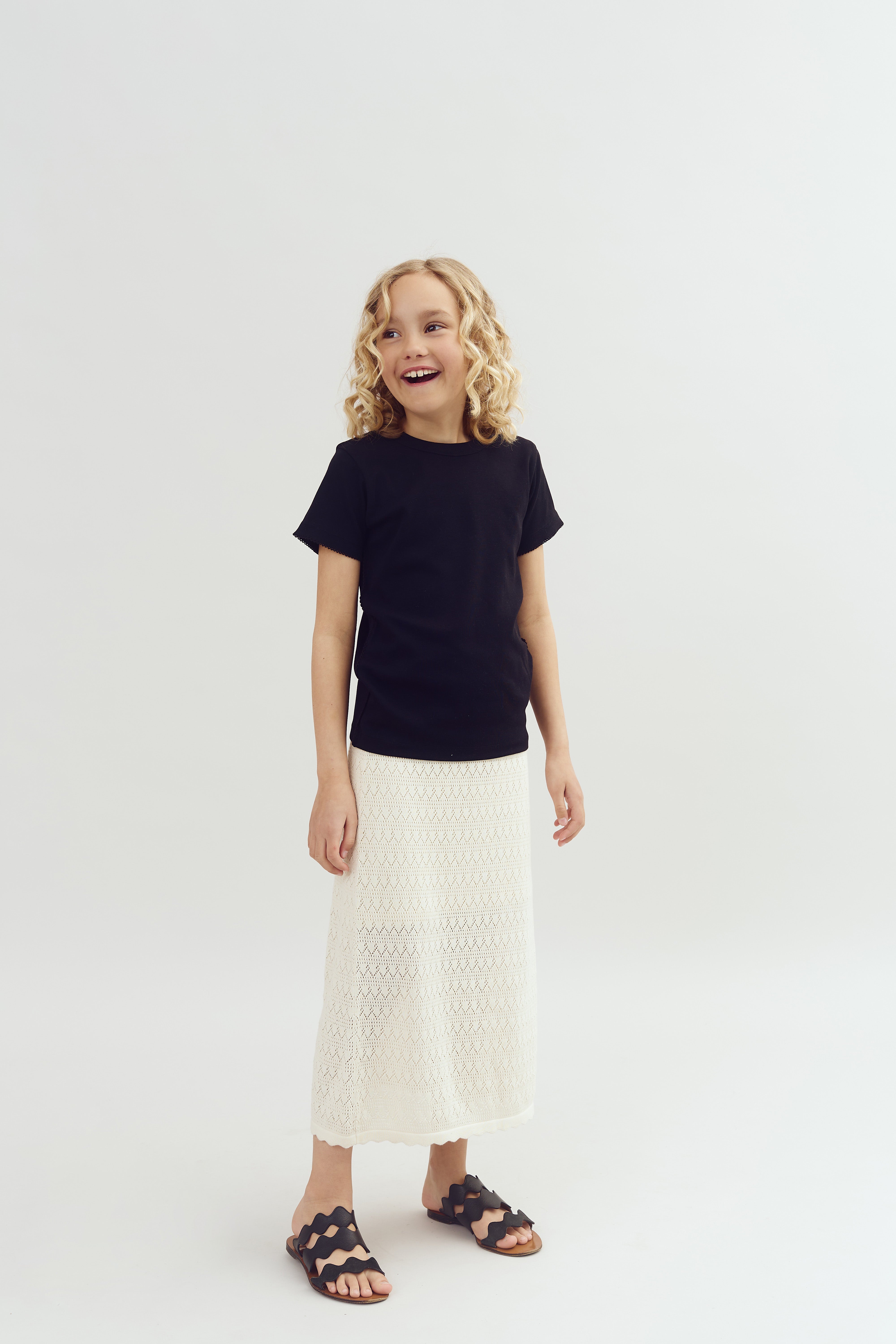 A child stands smiling, wearing a black shirt and a long white skirt, against a plain white background, exuding a cheerful demeanor.