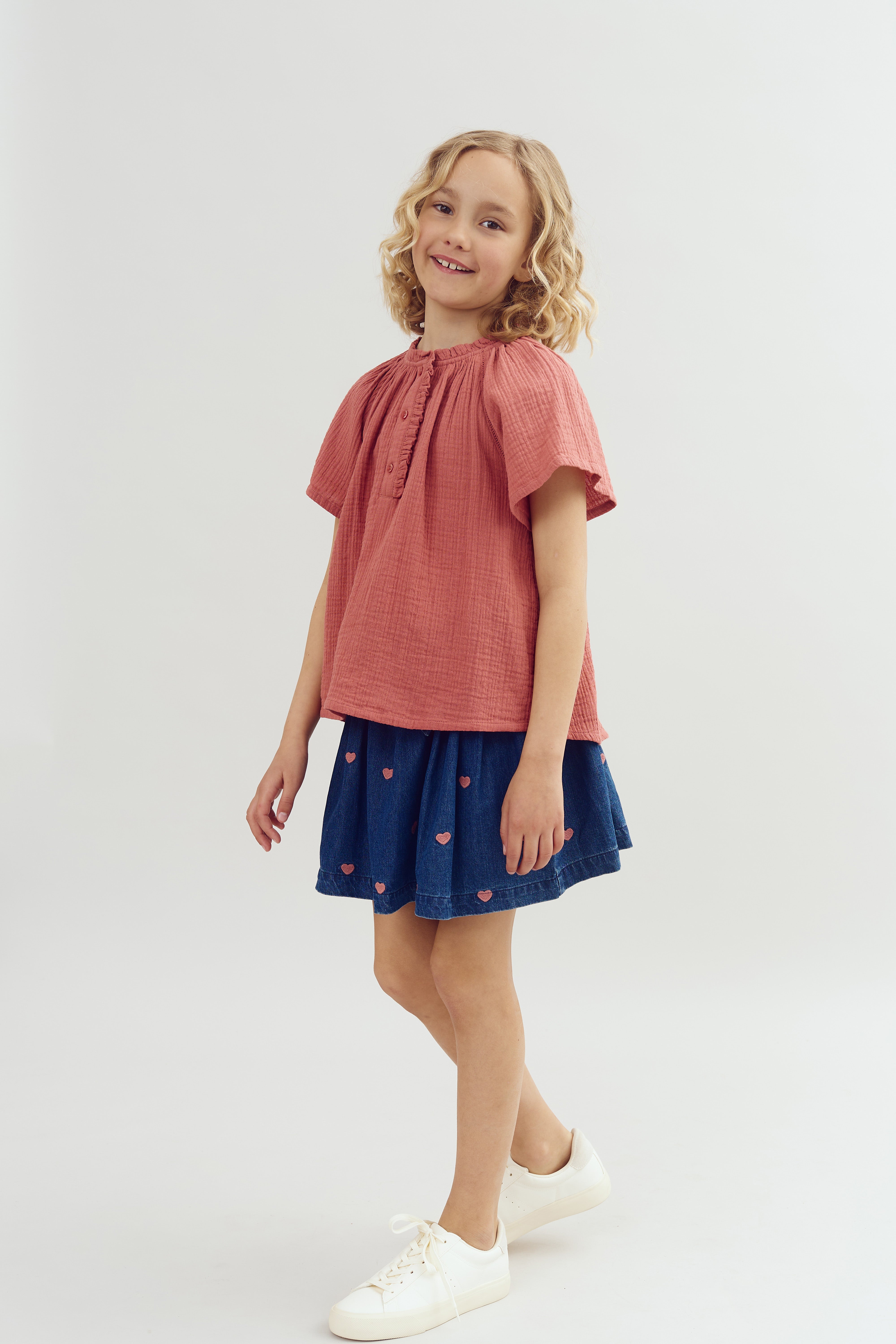 A child stands smiling, wearing a pink top and a blue skirt with heart patterns, in a neutral studio setting.