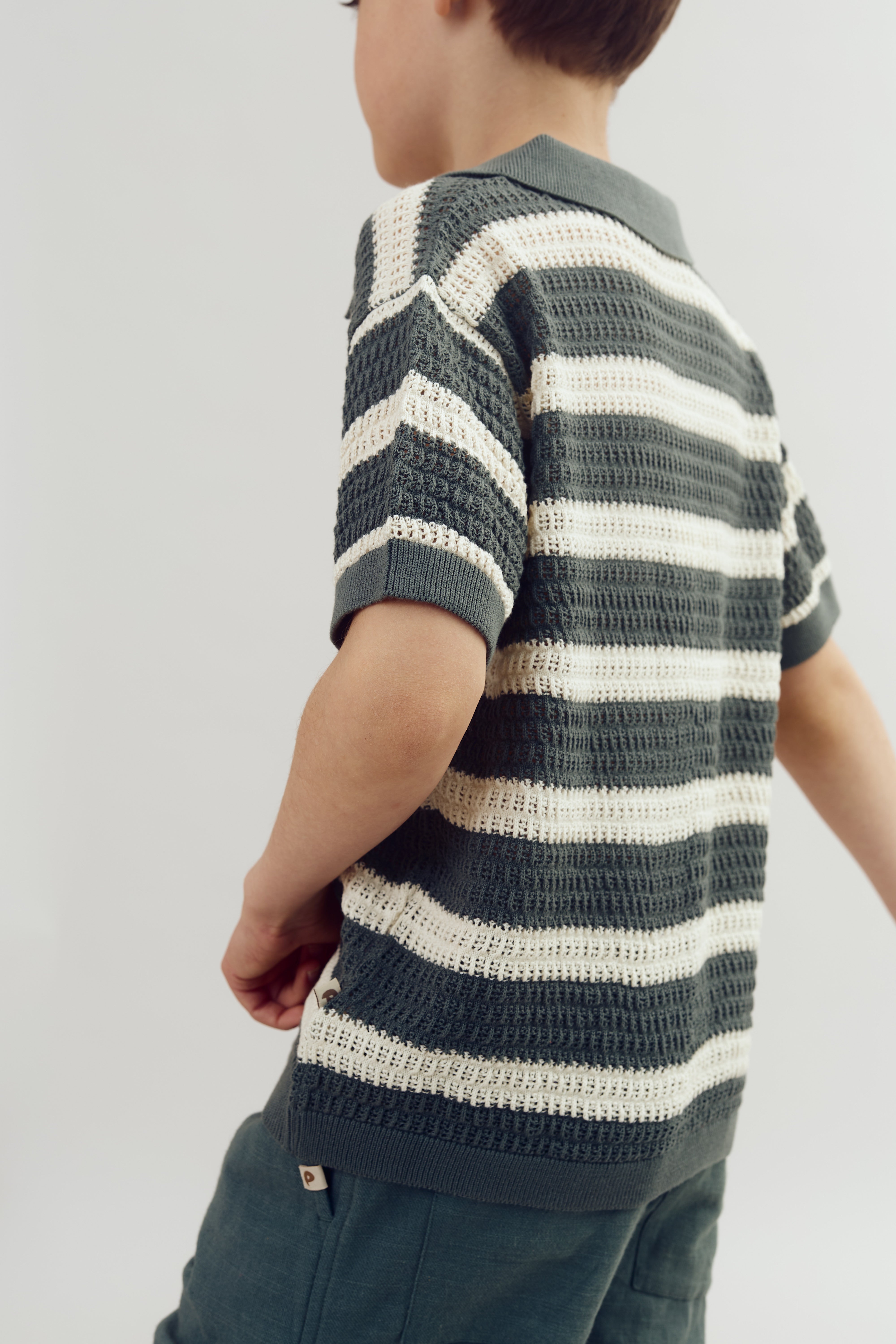 A child wears a green and white striped knit shirt, standing sideways against a plain background, with green pants visible below the waist.