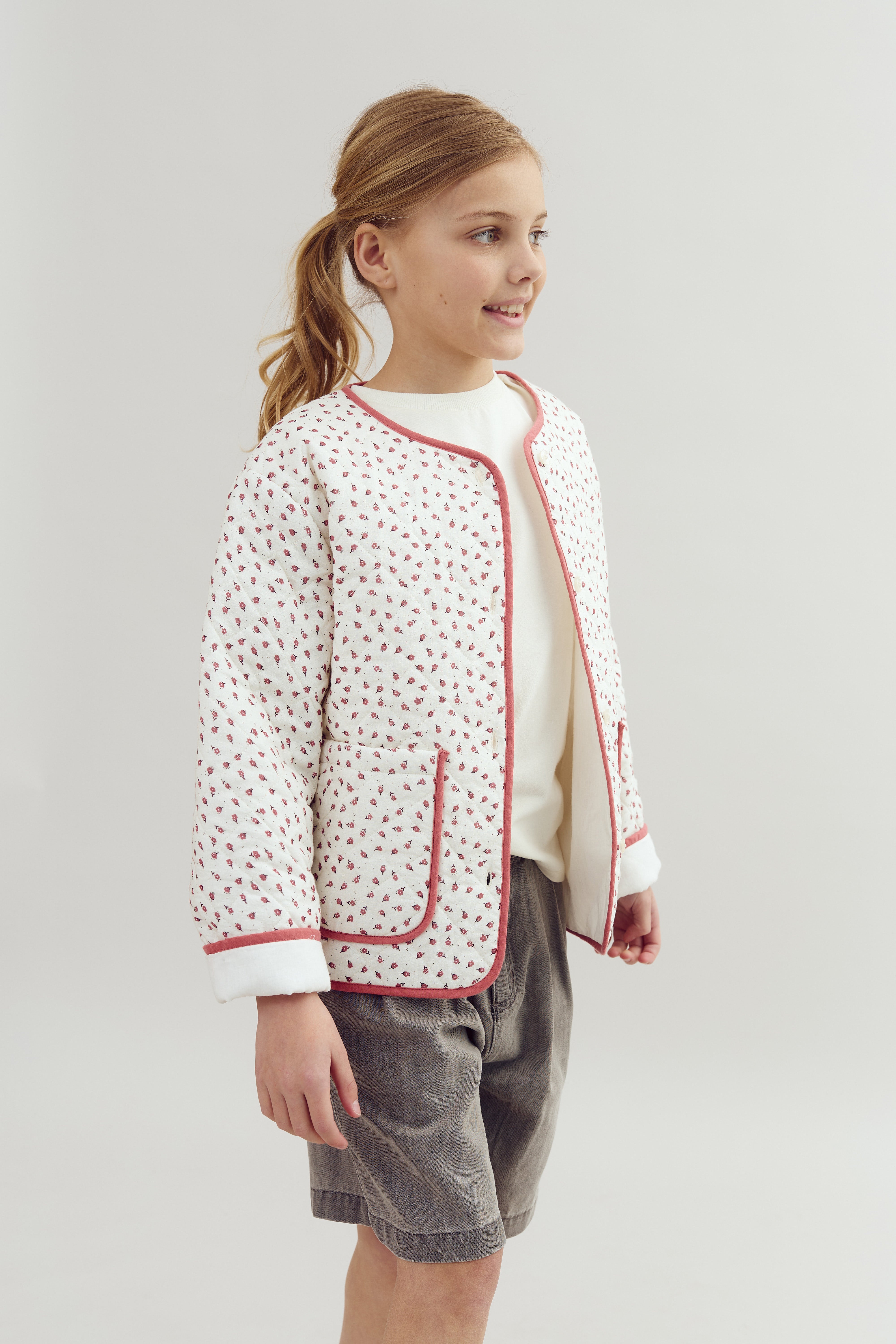 A child wearing a patterned jacket with red trim stands smiling in a minimalist, well-lit space with a plain background.
